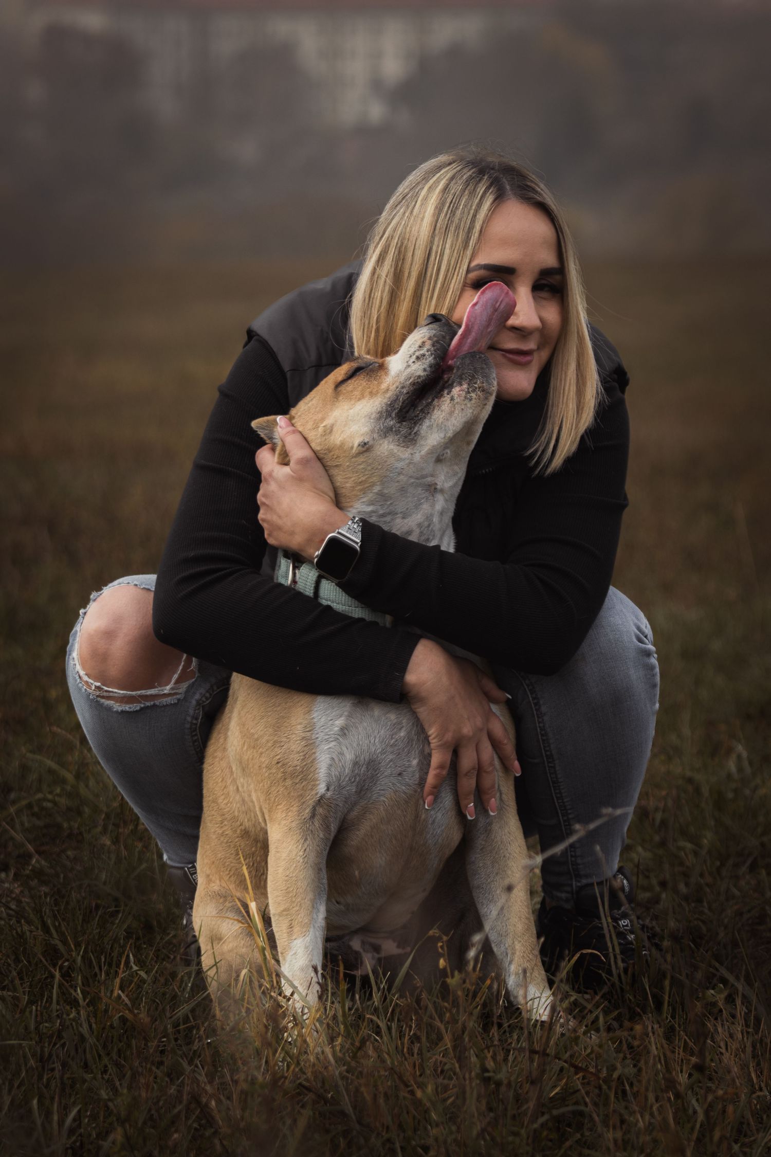 szőke nő fekete pulcsiban és szakadt farmerben guggolva ölel egy kutyát, ami megnyalja az arcát