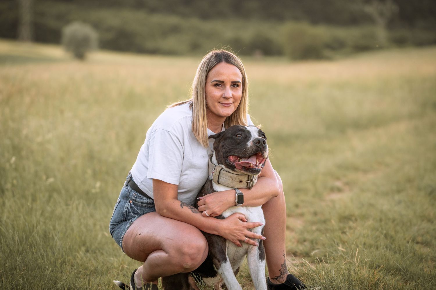 Woman in white tee and denim shorts crouching with a brown and white dog in a sunny green field.