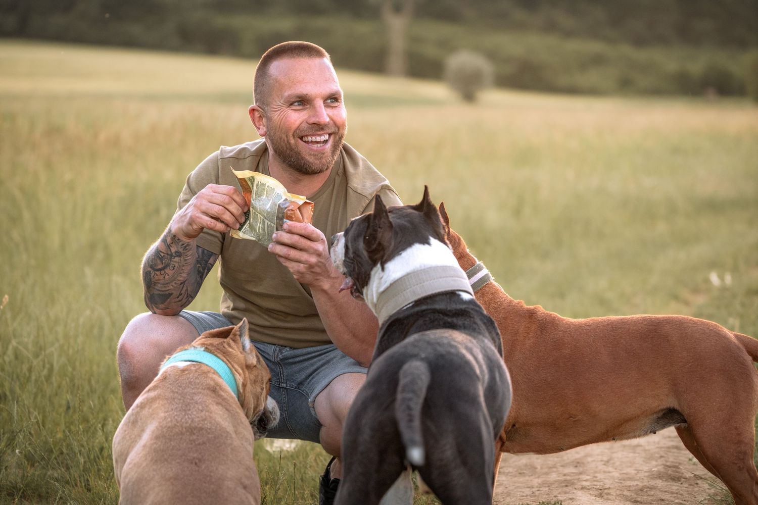 Tattooed man smiling outdoors in a field, crouching beside two boxer dogs while holding a treat.