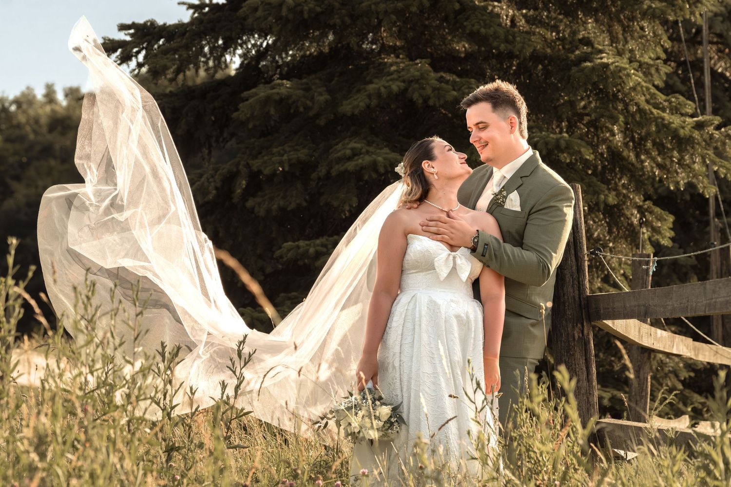 Bride and groom embrace in golden sunlight with wedding veil flowing in the wind against a rustic outdoor backdrop.