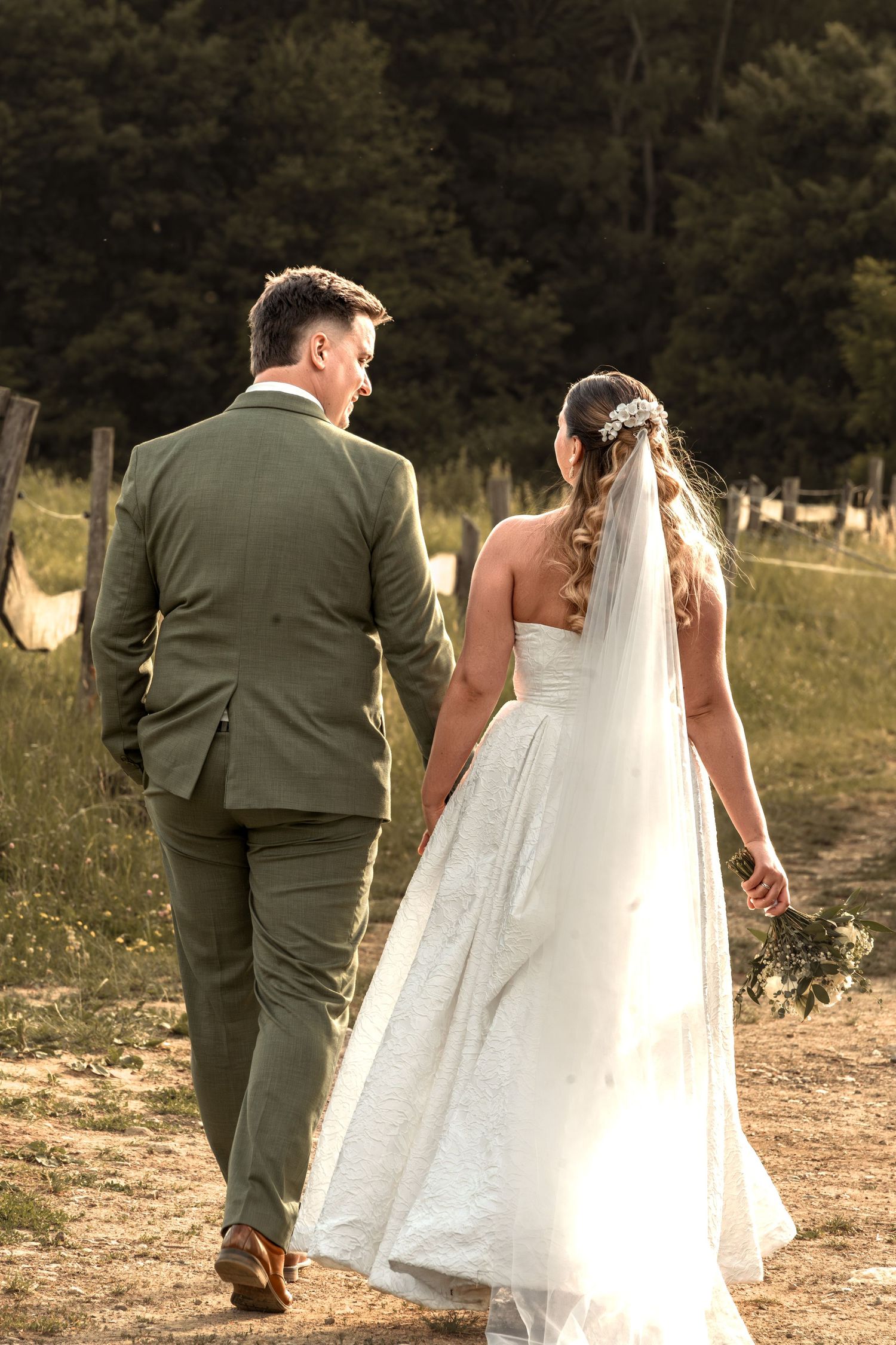 Bride and groom walking hand in hand through a sunlit field, groom in green suit, bride in white lace gown with veil.