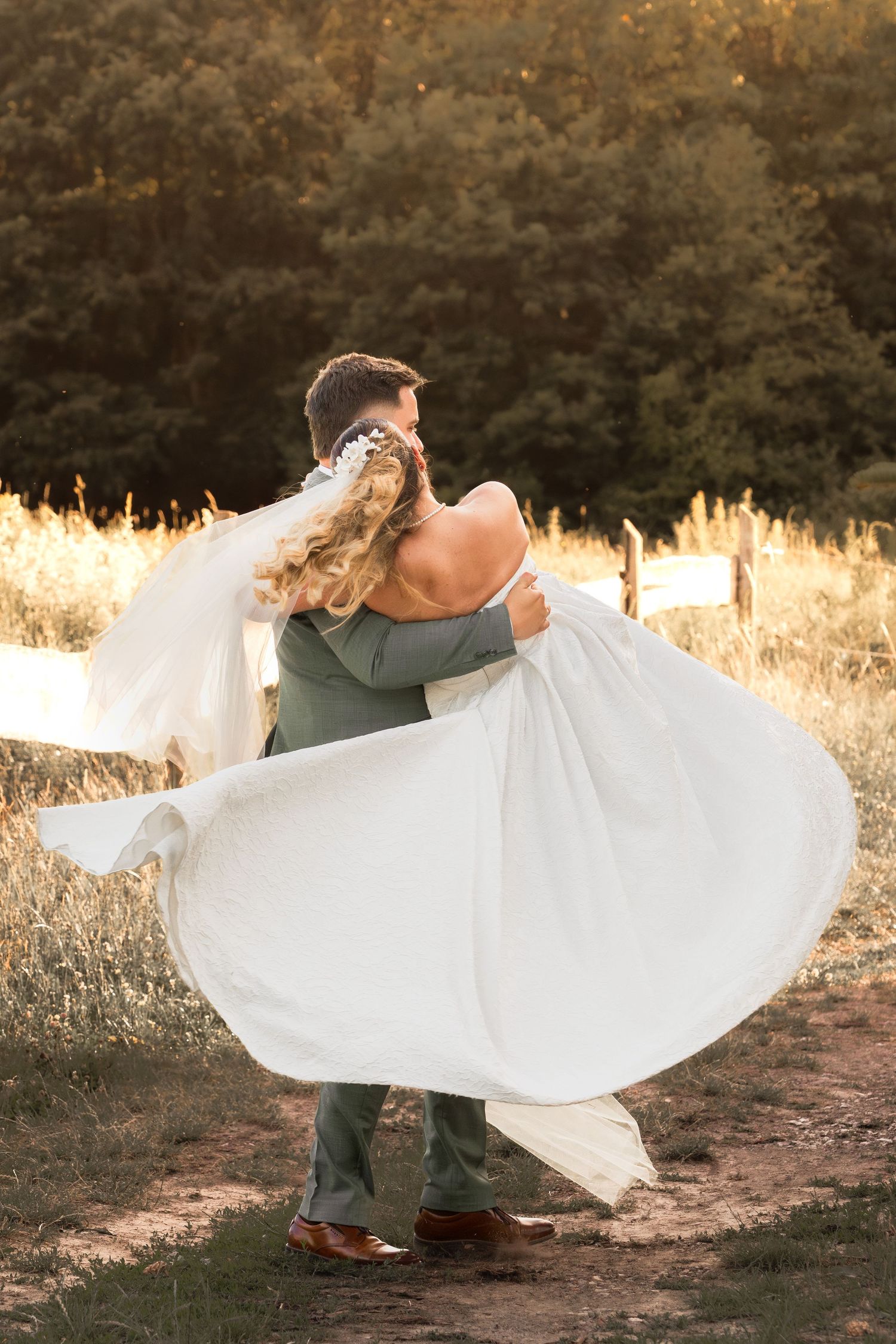 Groom lifting bride in white flowing dress outdoors at golden hour, surrounded by lush greenery.