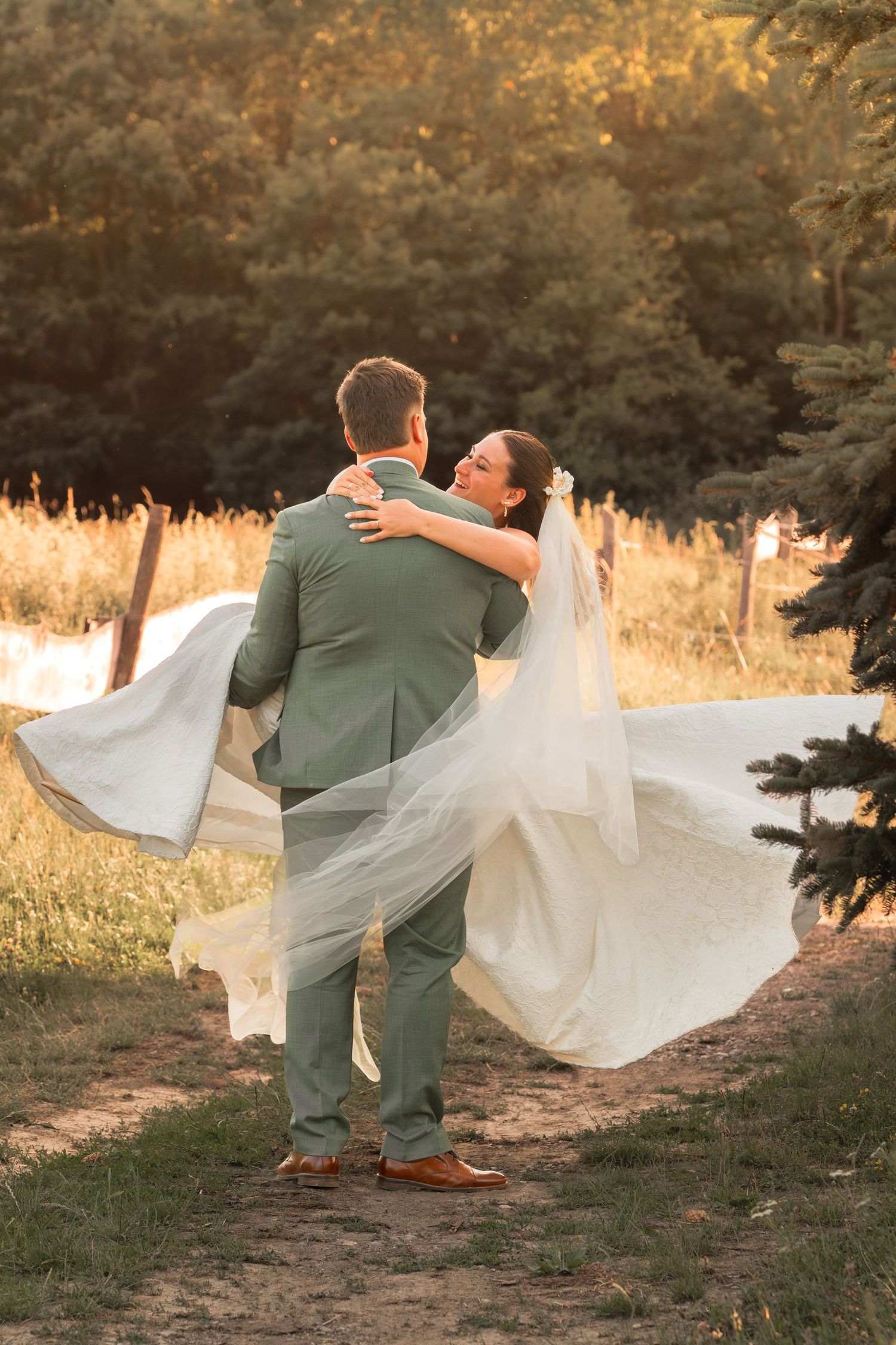 Groom in sage green suit lifts bride in flowing white wedding dress and veil outdoors at golden hour.