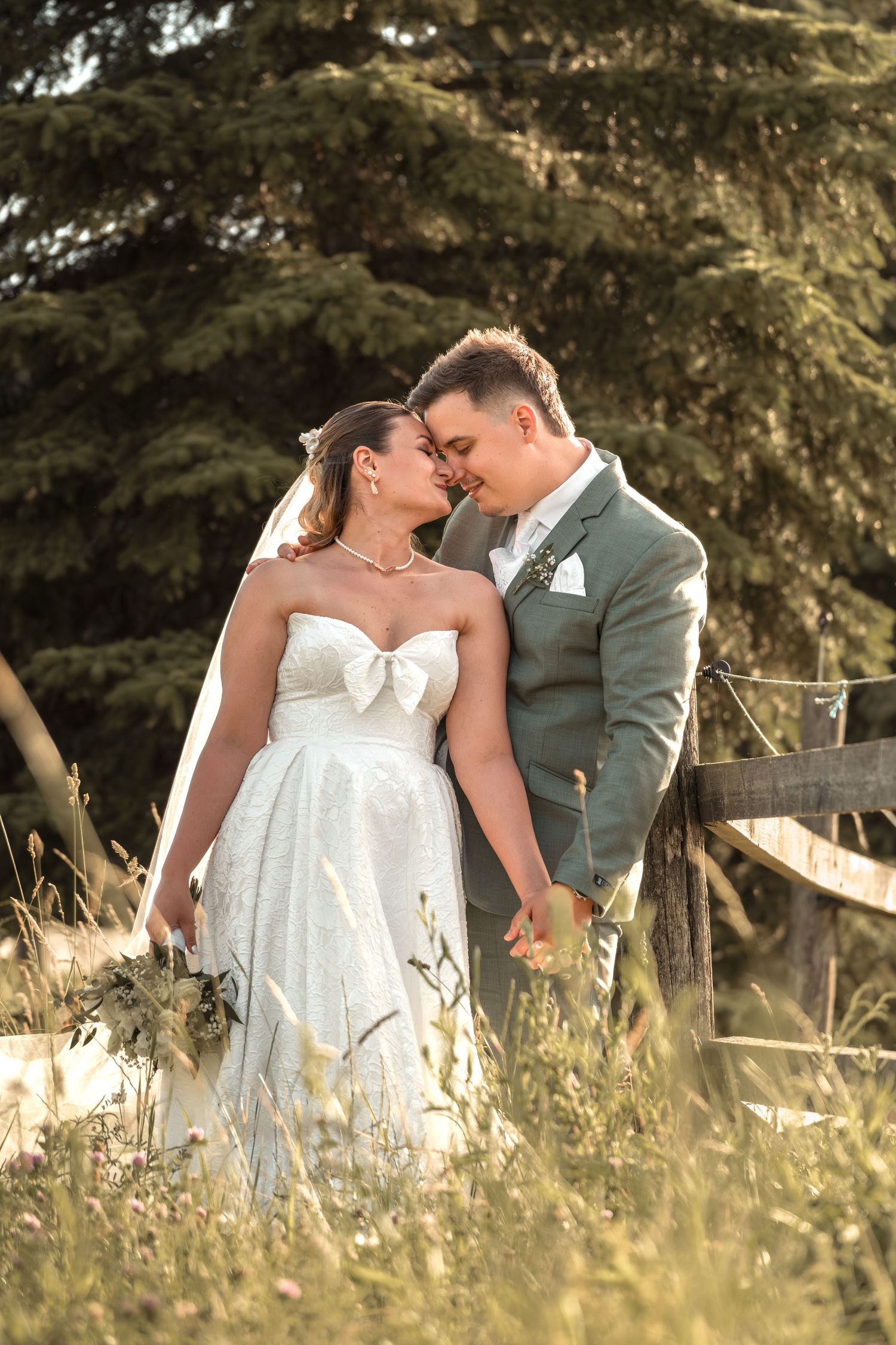 Bride and groom share romantic moment in field, she in strapless white gown holding bouquet, he in sage suit.