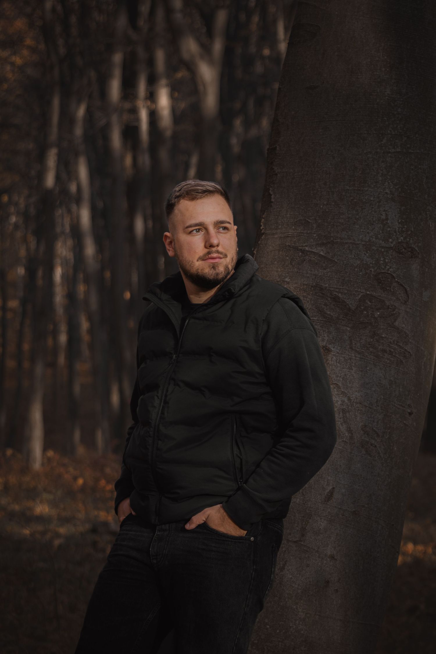 fiatal férfi fekete kabátban fának támaszkodva egy sötétebb őszi erdőben