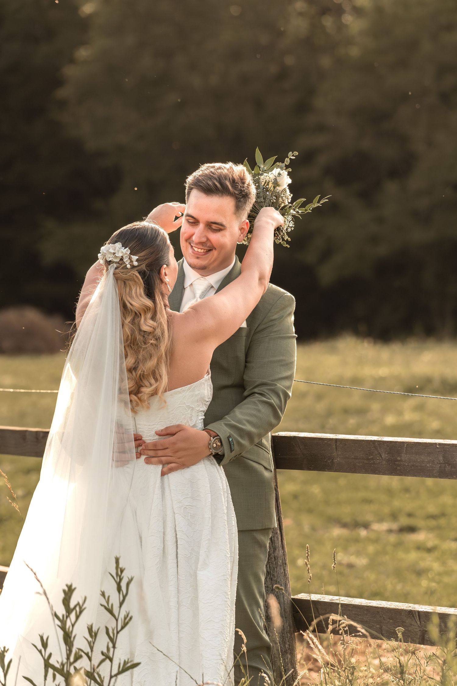 Bride and groom embracing outdoors at golden hour, smiling joyfully, bride holding bouquet in rustic field setting.