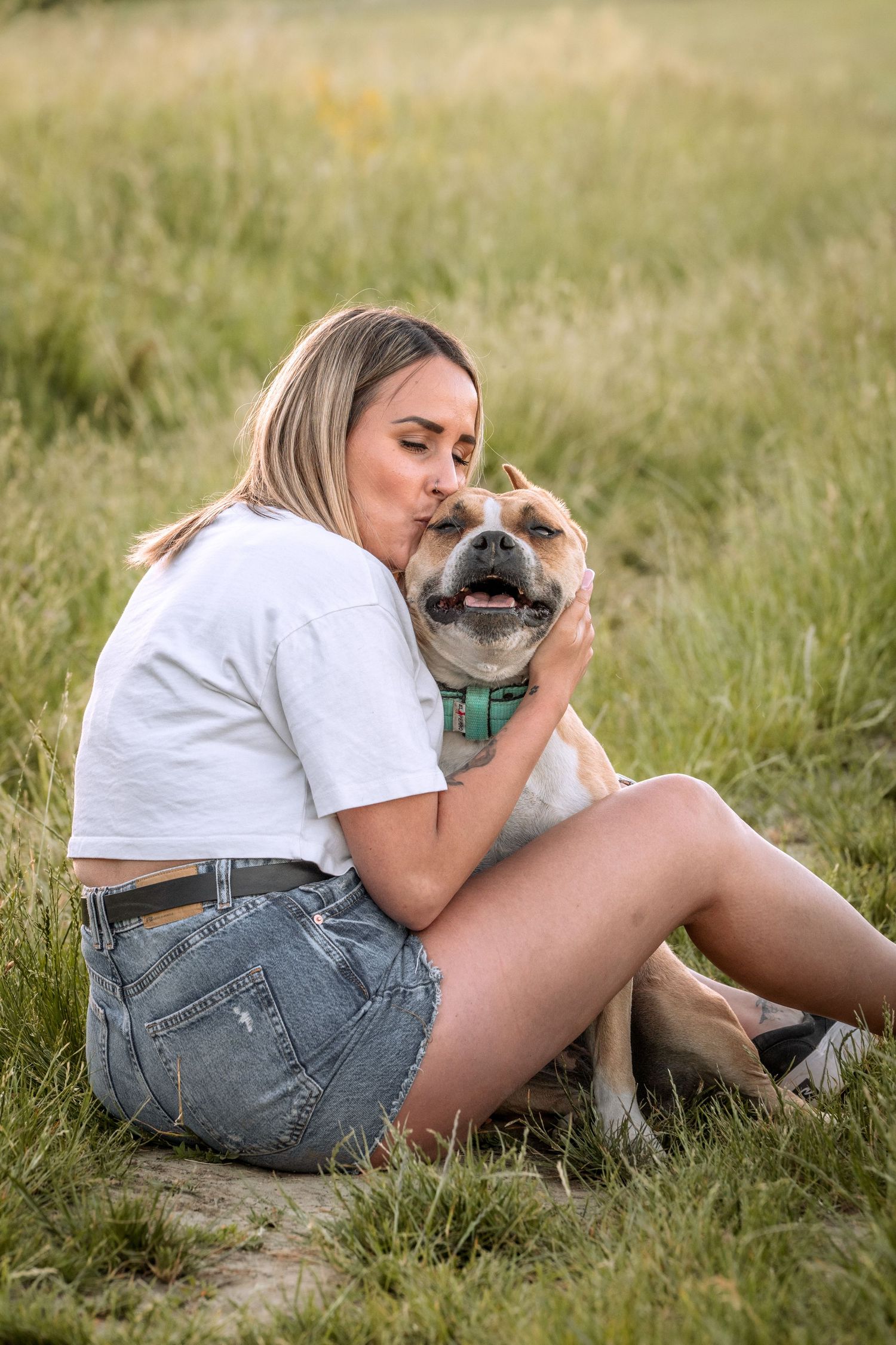A young woman in white tee and denim shorts sits in a grassy field, lovingly kissing her tan dog wearing a green collar.