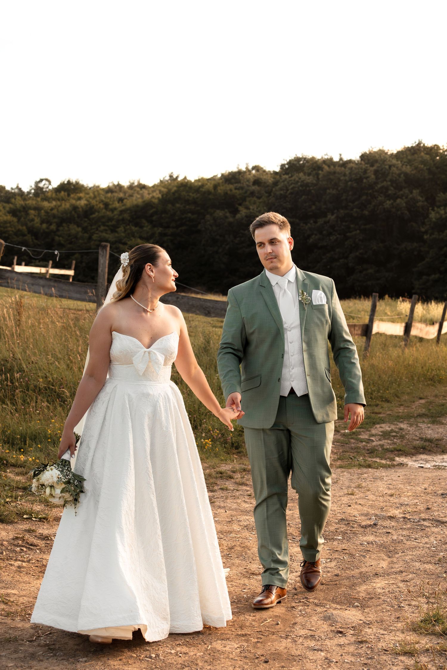 Bride in white strapless gown and groom in sage green suit walk hand in hand on a dirt path at golden hour.