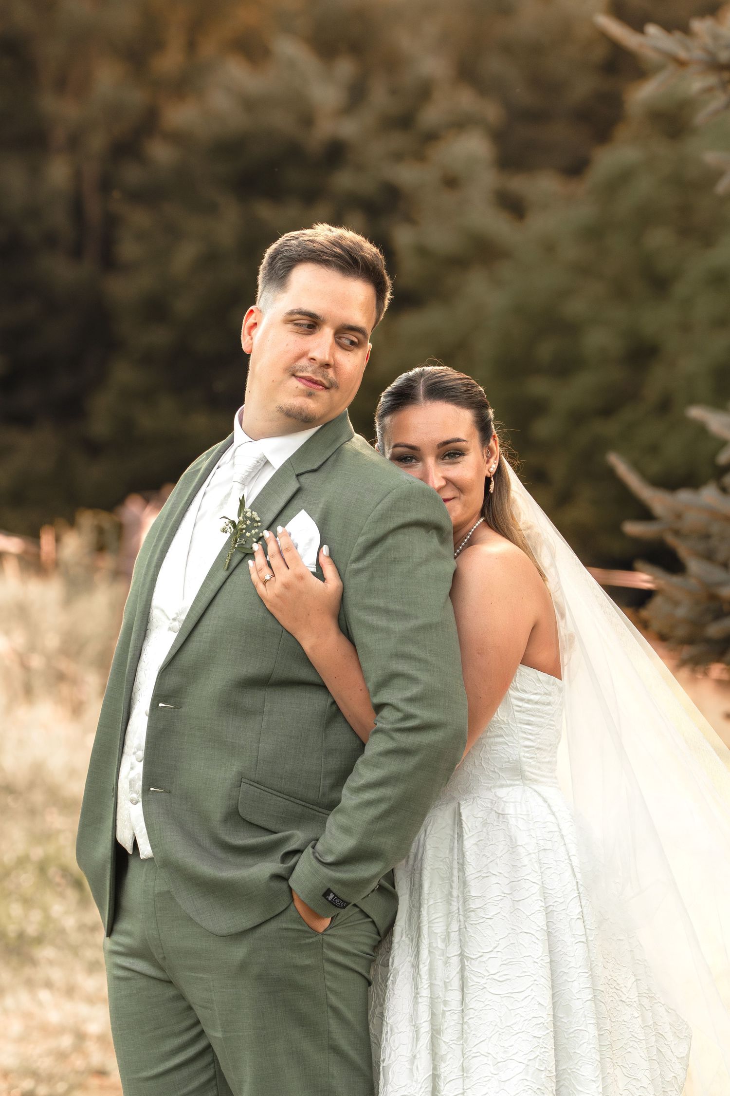Bride in white gown and veil leans on groom in sage green suit with boutonniere, posing outdoors.