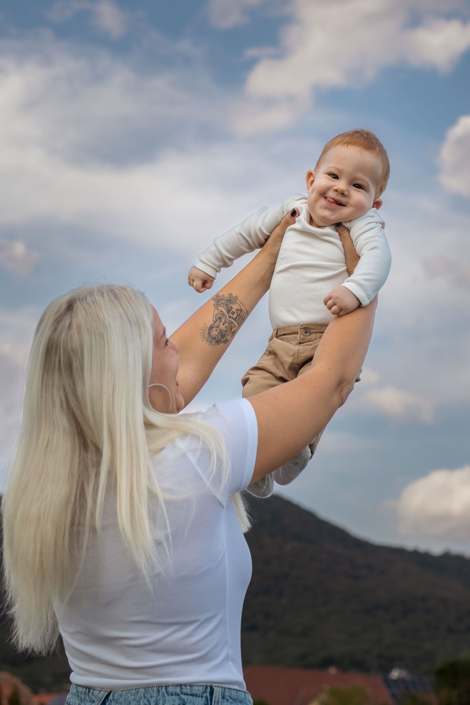 szőke anya a magasba emeli mosolygó kisfiát a szabadban