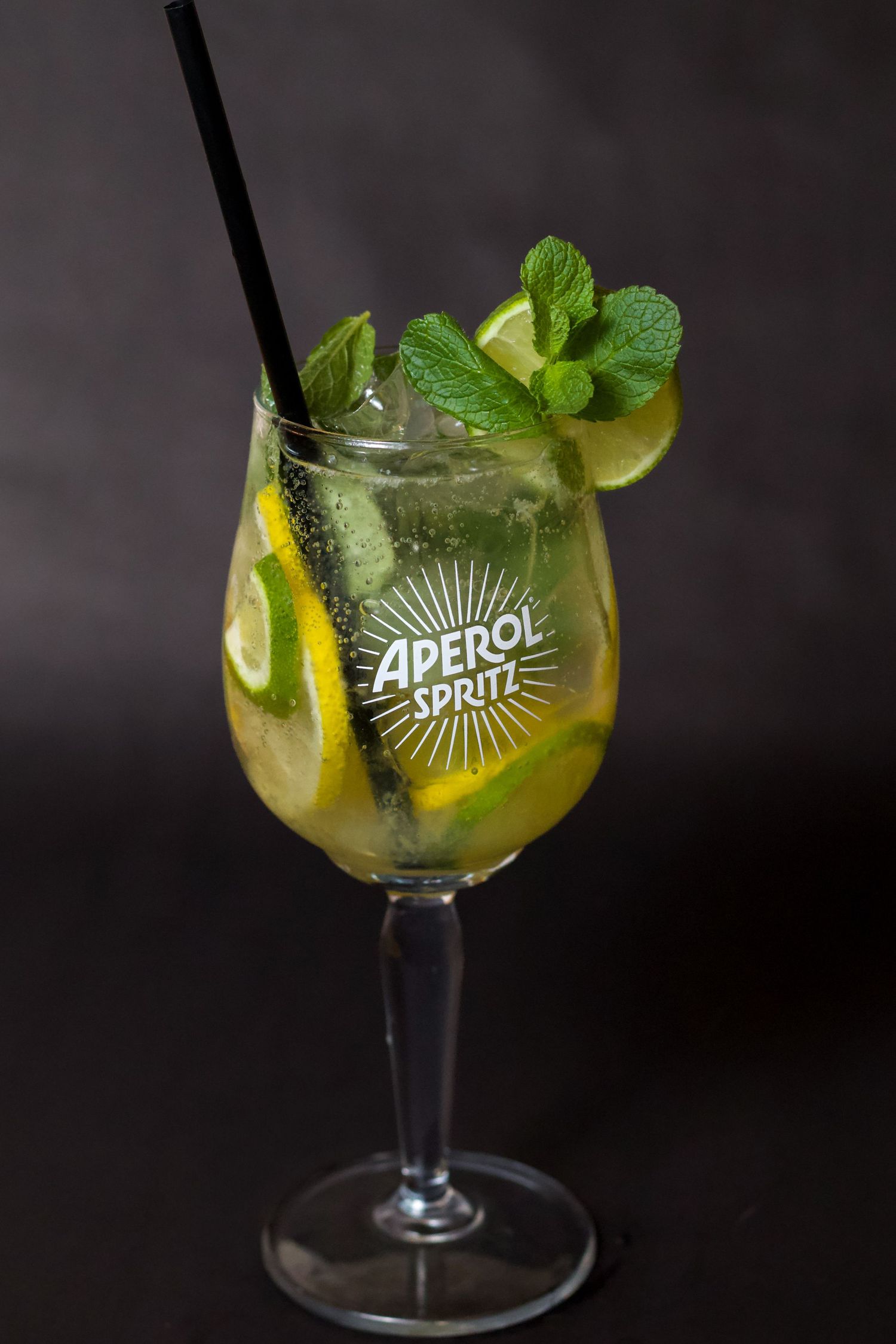 Hugo Spritz koktél márkázott pohárban, mentával, lime-mal és jéggel sötét háttér előtt