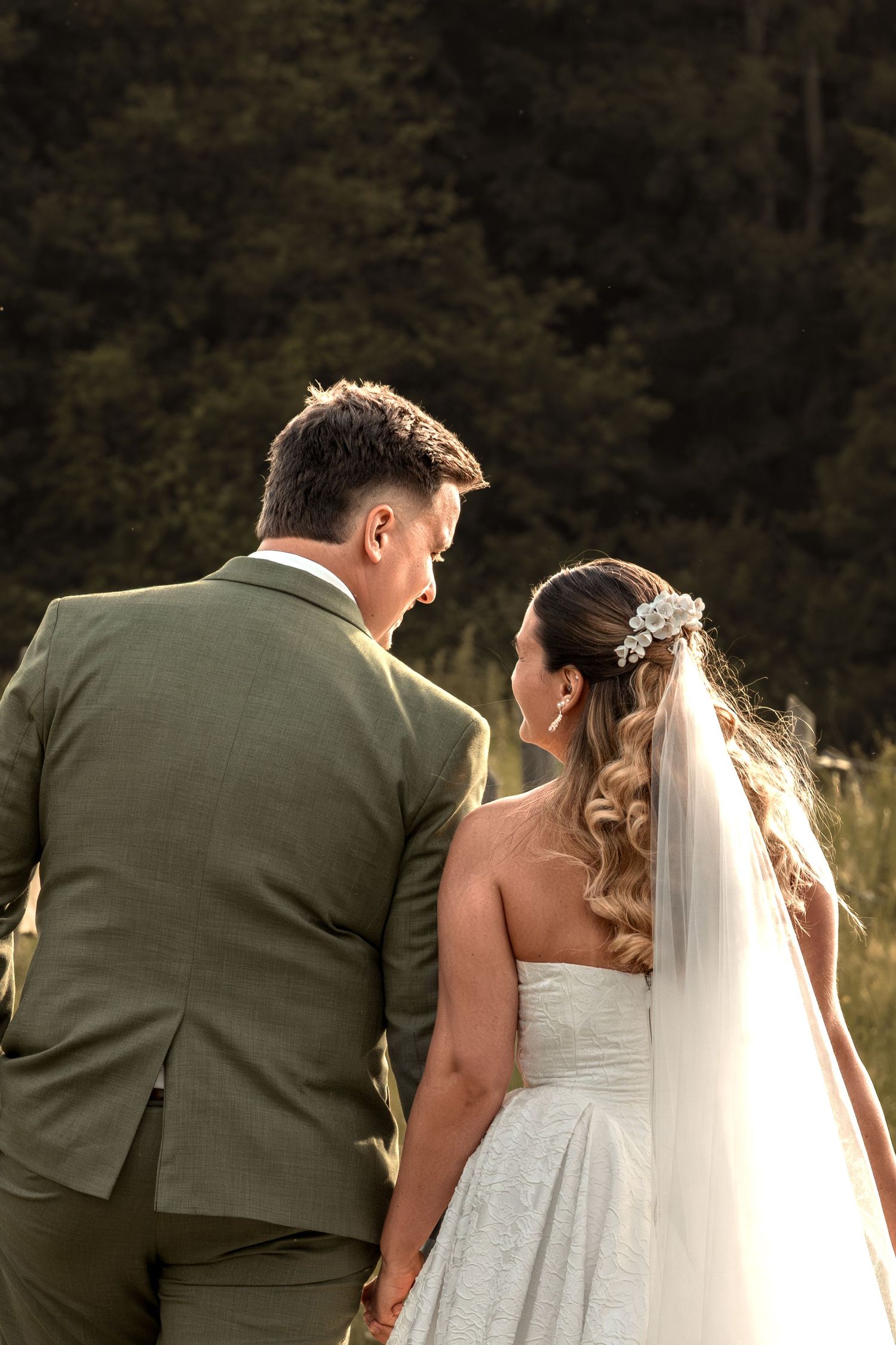 A bride and groom hold hands walking through a sunlit field, she in a white gown with veil, he in a sage green suit.