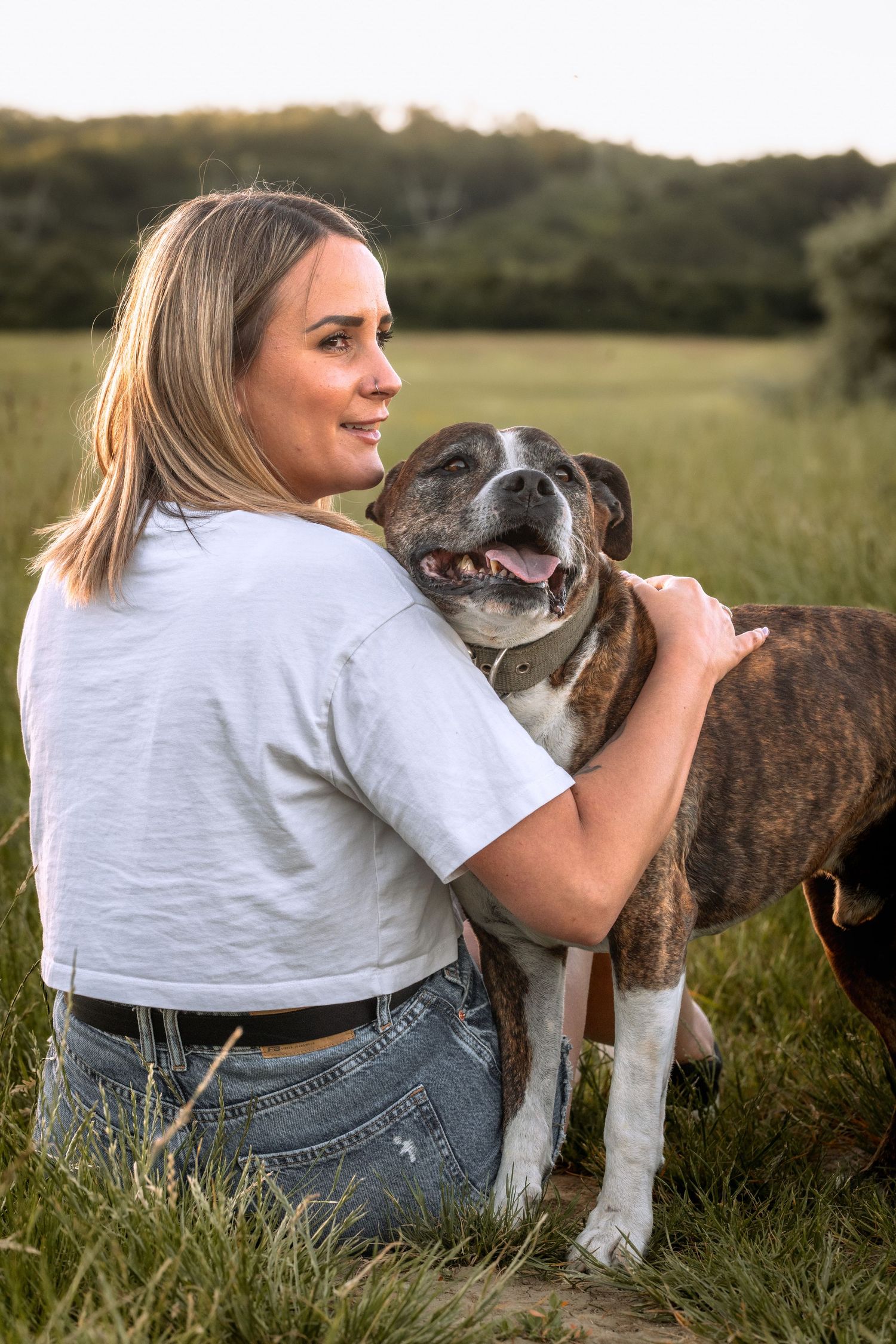 Woman sitting in a green field hugging her brindle Boxer dog during golden hour sunset.