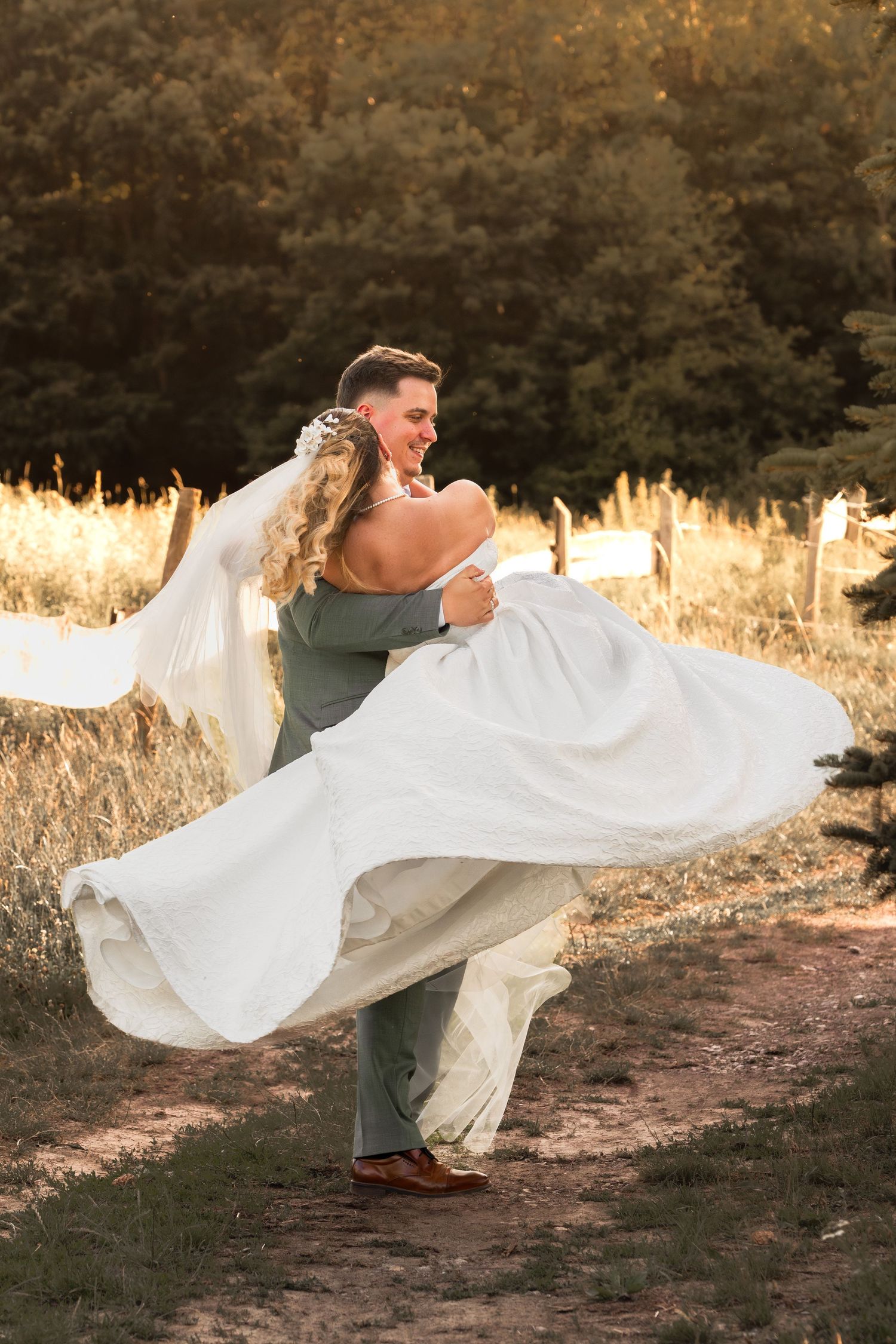 Groom lifting bride in white gown, spinning joyfully on a sunlit path surrounded by golden fields and lush green trees.