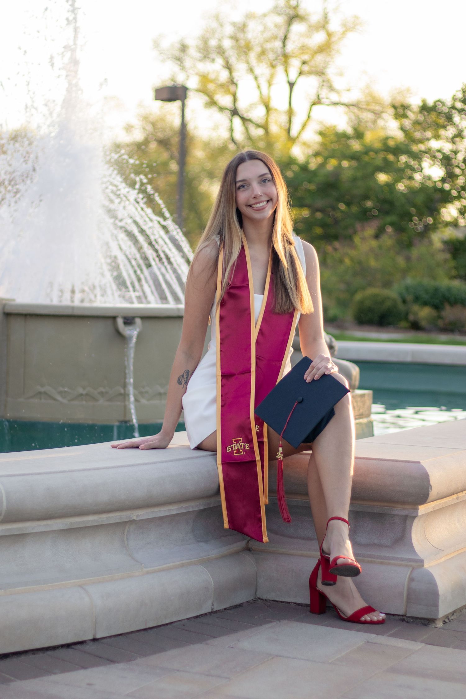 Smiling female graduate in white dress and maroon stole posing by a fountain.