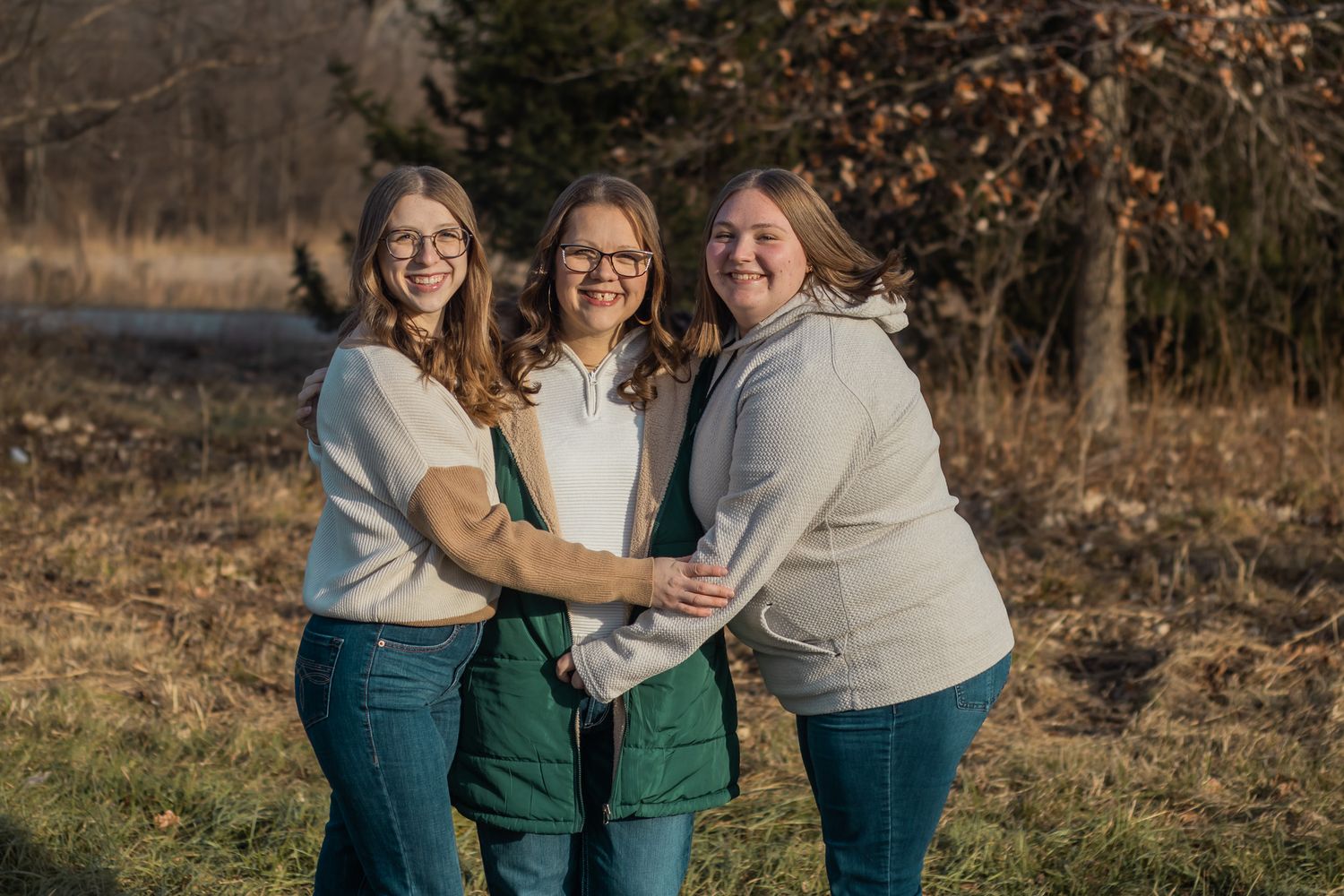 Three women smiling and embracing outdoors in warm natural light.