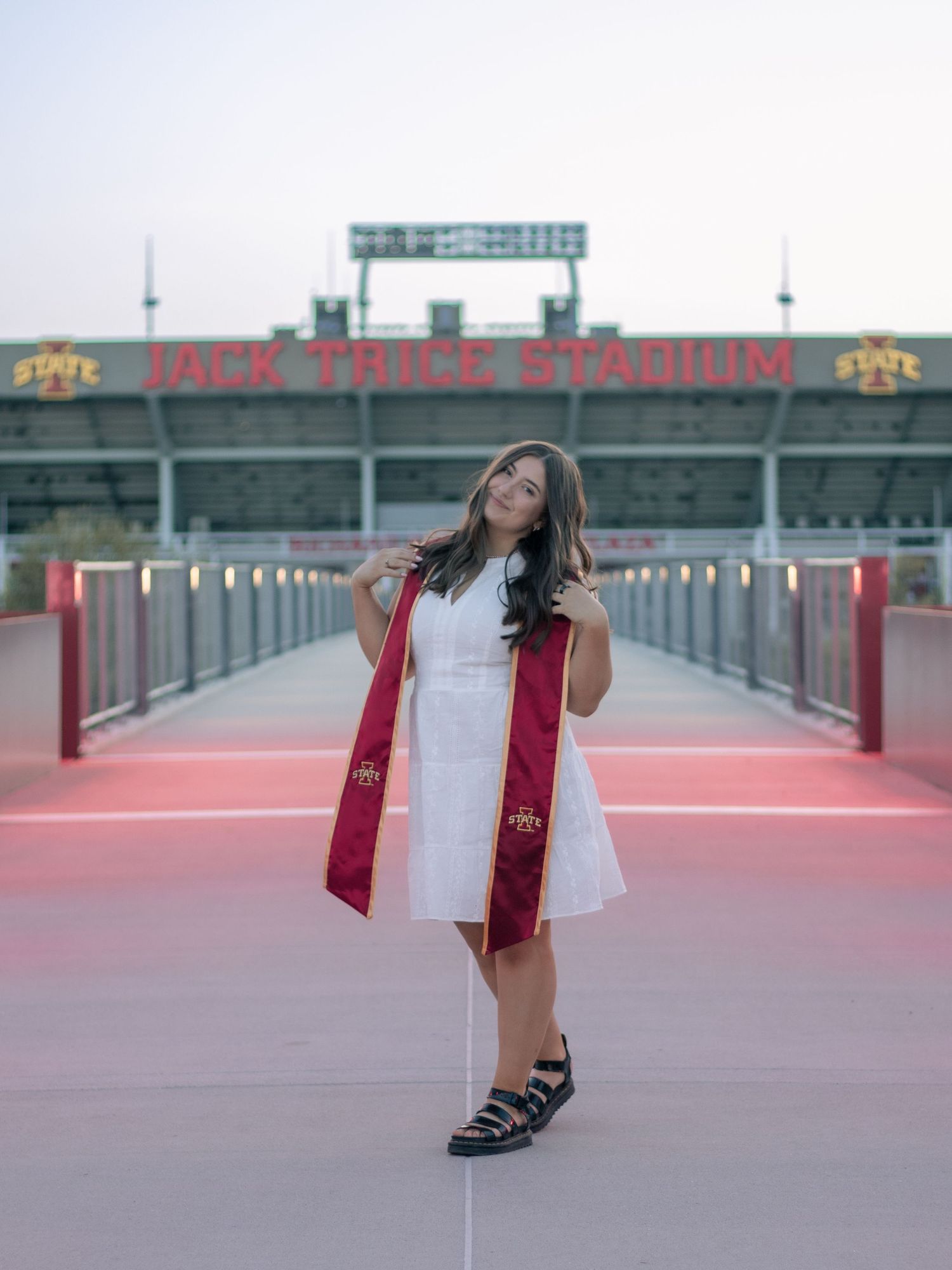 Iowa State University graduate poses in white dress with cardinal stole at Jack Trice Stadium during golden hour.