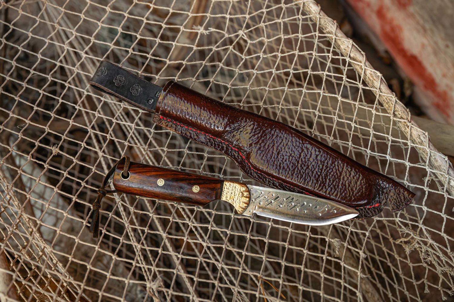 Käsintehty puukko puisella kahvalla ruskean nahkatupen vieressä kalaverkko taustalla.