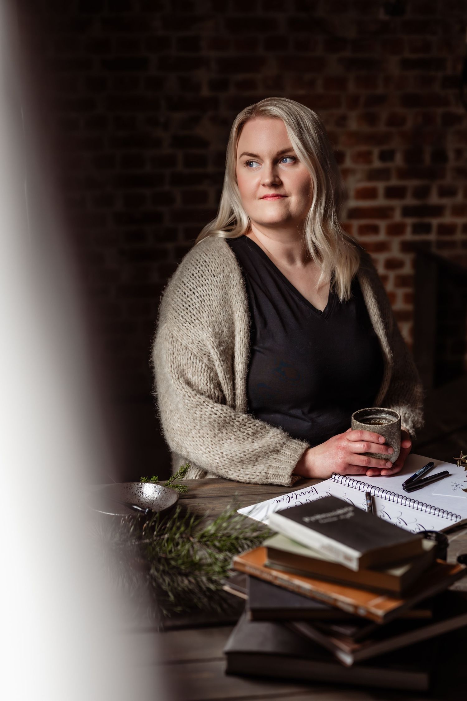 Vaaleahiuksinen nainen beigenvärisessä neuletakissa juoma kädessään, istuu työpöydän, jossa kirjoja ja kirjoitusvälineitä, ääressä.