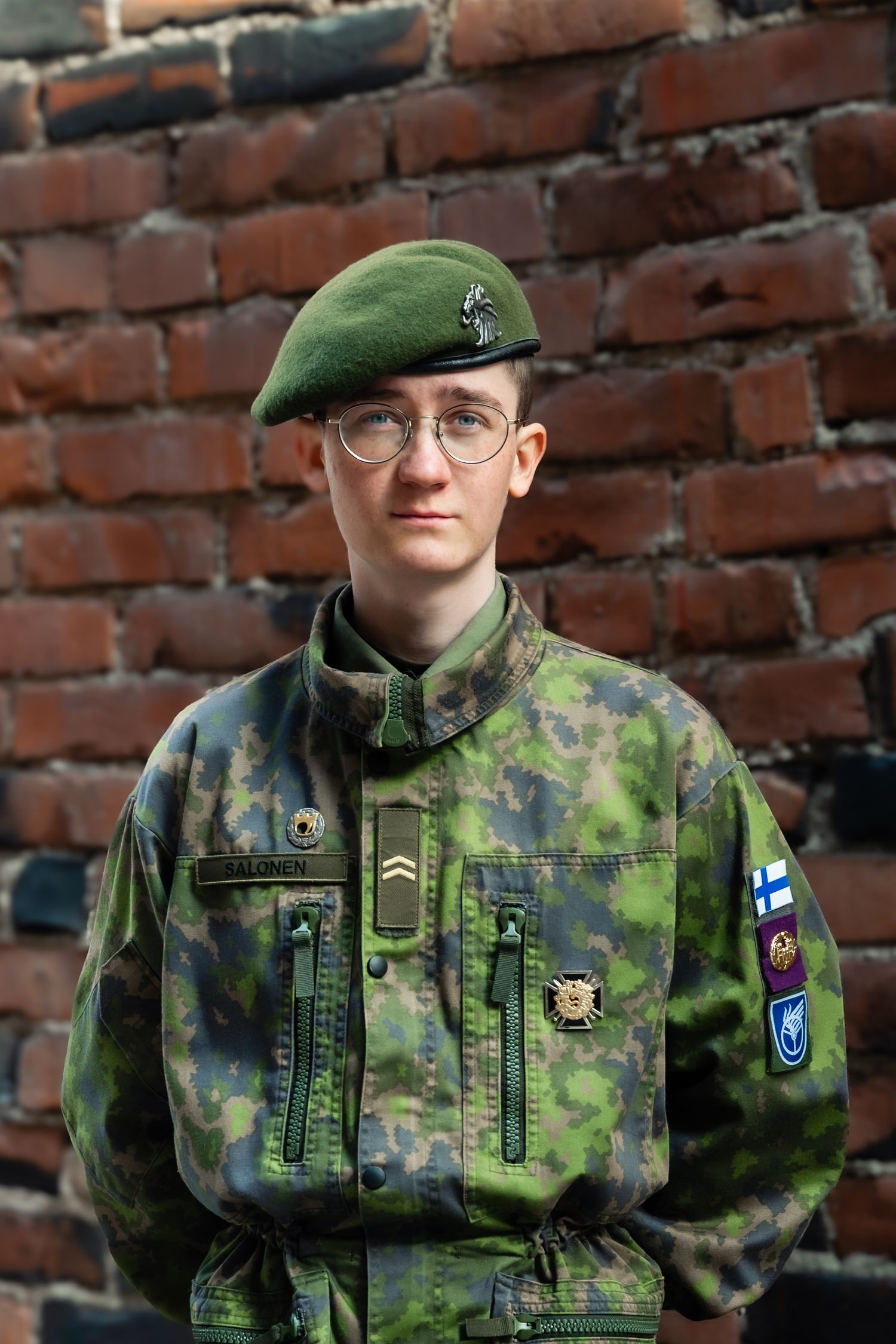 Nuori suomalainen sotamies vihreässä armeijan asussa ja baretissa seisoo tiiliseinän edessä.
