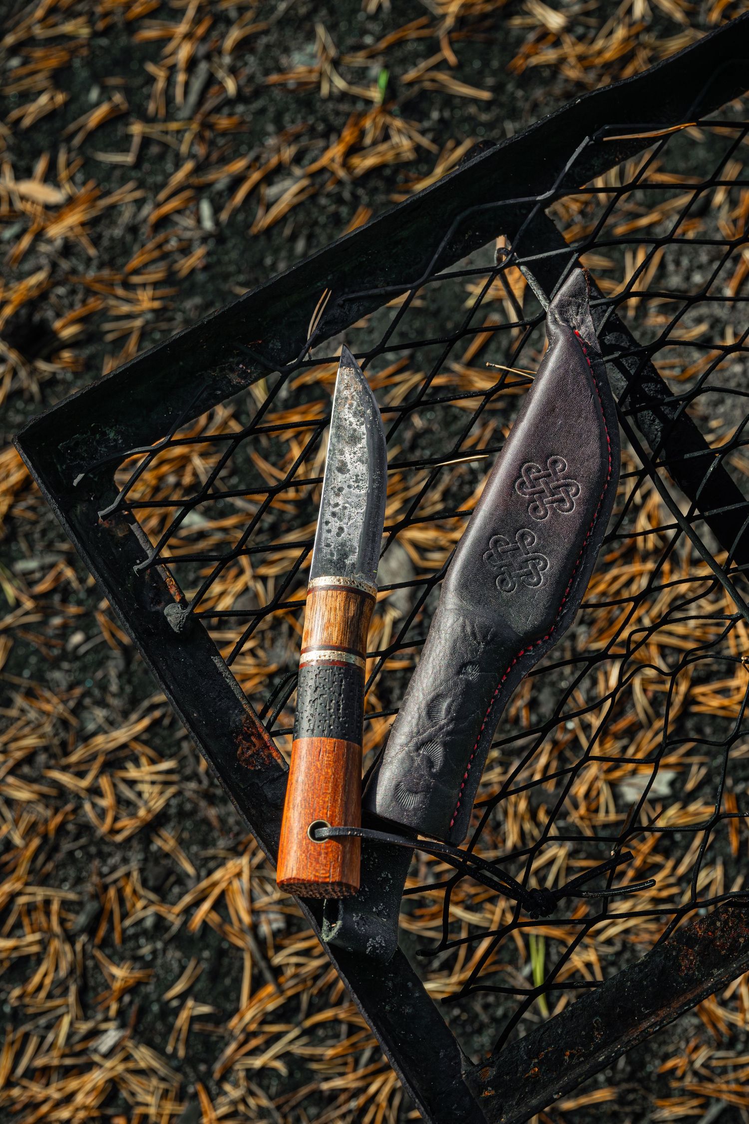 Käsintehty puukko puisella kahvalla kaiverretun nahkatupen vieressä metalligrillin ritilällä ulkona.