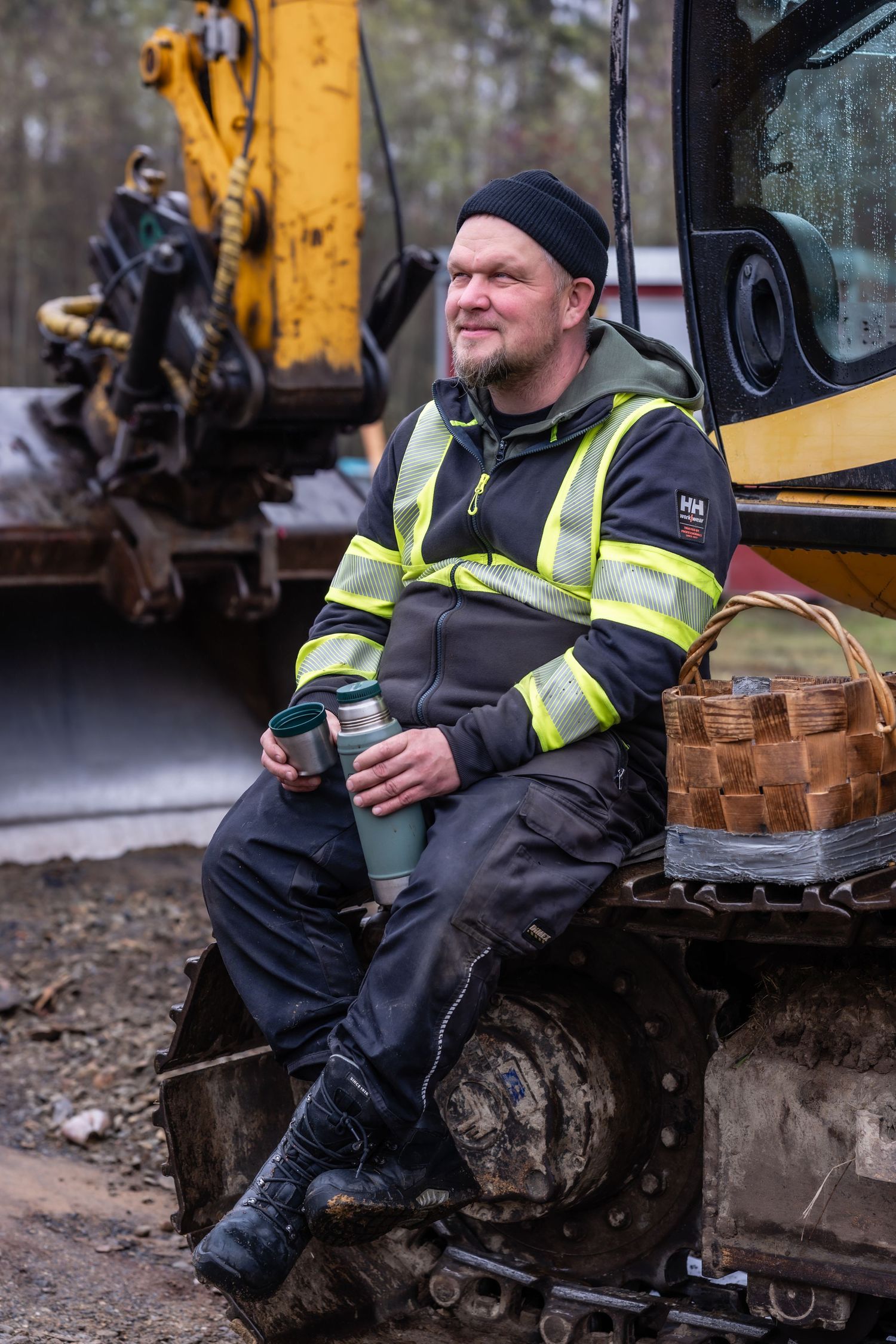 Ammattilainen heijastintakissa istuu kaivinkoneen äärellä termospullo kädessään tauon aikana ulkona.