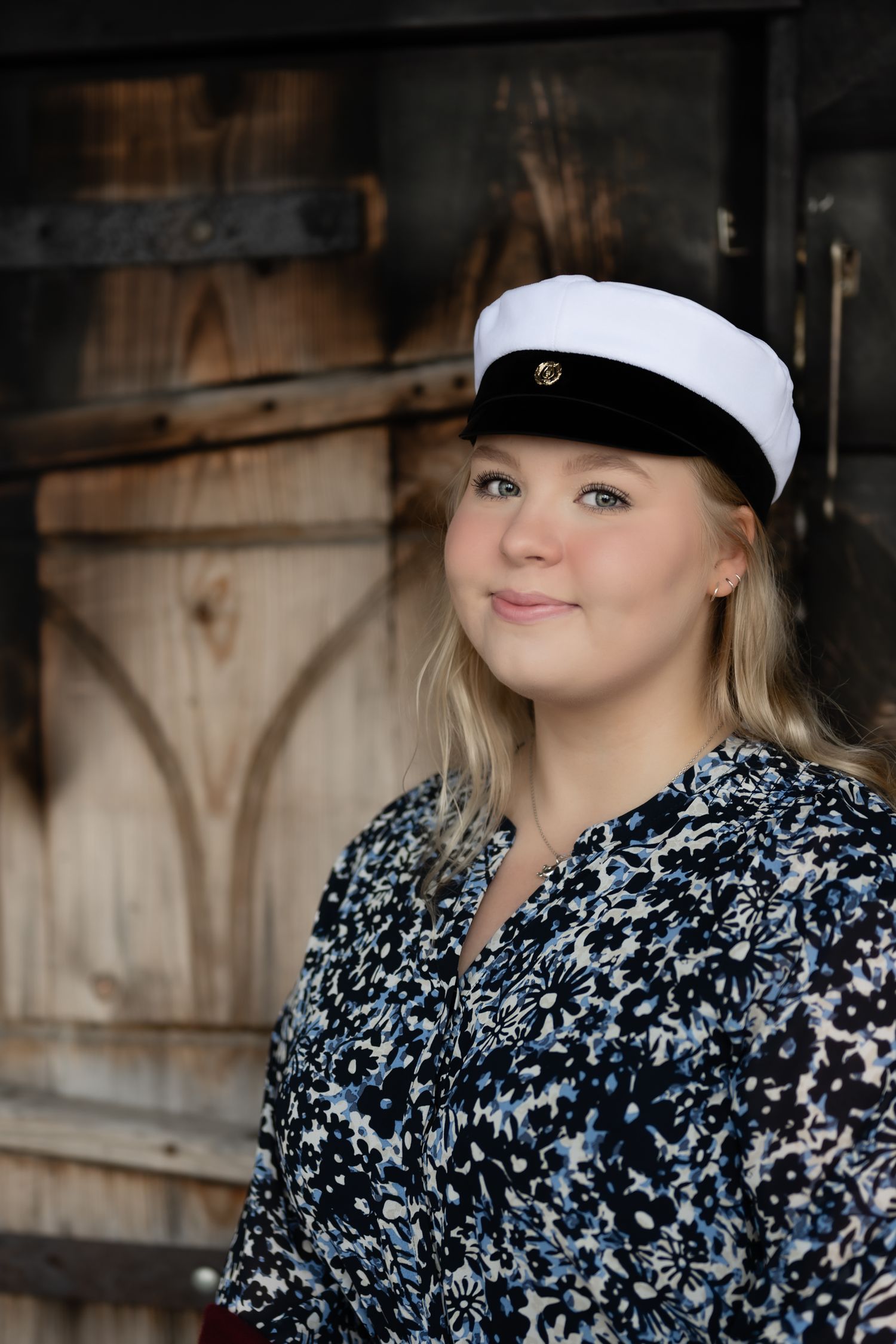 Nuori vaalea nainen kukkakuvioisessa mekossa ja ylioppilashatussa poseeraa maalaismaisen puisen oven edessä.