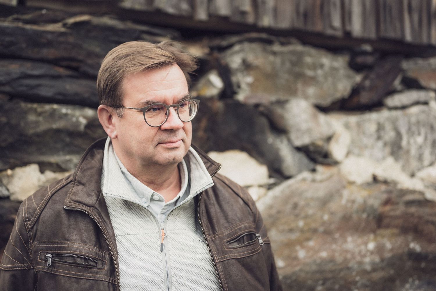 Keski-ikäinen mies silmälaseissa ja ruskeassa nahkatakissa seisoo ulkona rakennuksen kiviperustusten edessä.