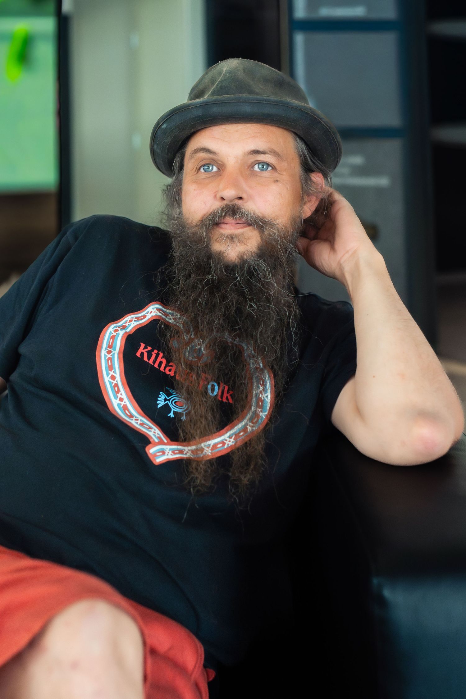 Parrakas ja sinisilmäinen mies, jolla on musta graafinen t-paita ja harmaa hattu, istuu rentoutuneena käsi pään yläpuolella.