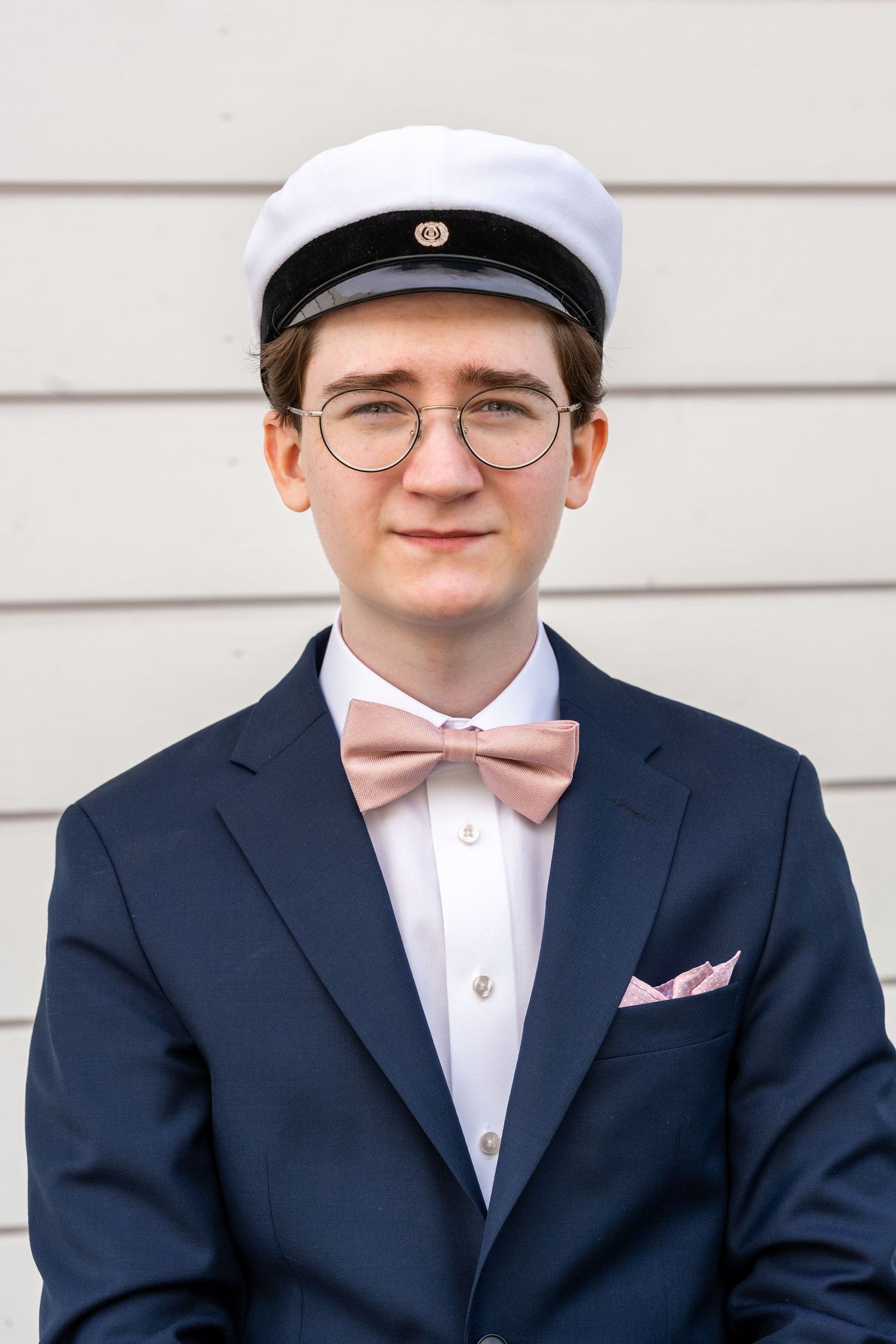 Tummansinisessä puvussa, vaaleanpunaisessa rusetissa, laseissa ja valkoisessa ylioppilaslakissa oleva nuori mies poseeraa muodollisesti.