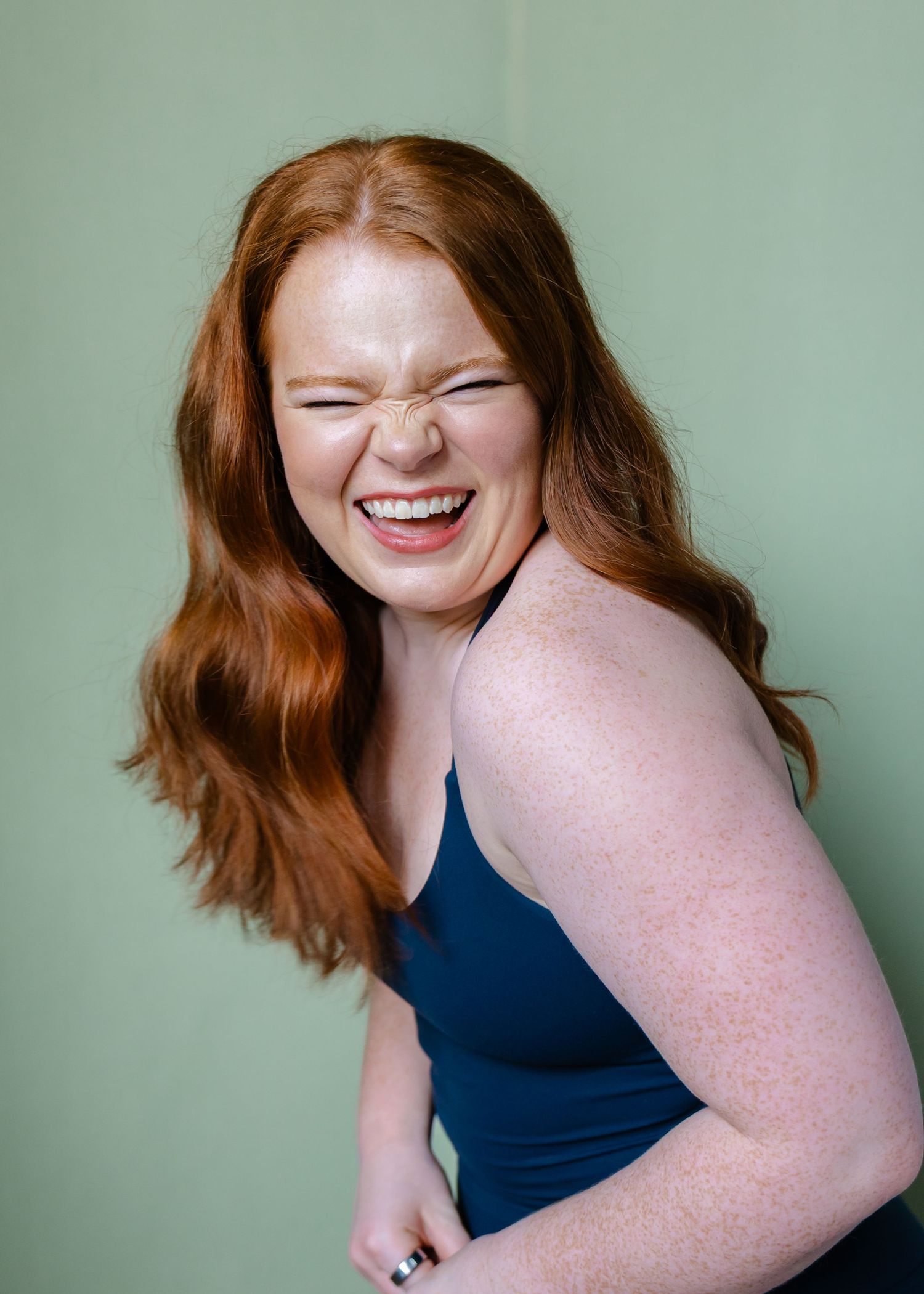 A red-haired freckled woman in a blue tank top laughs against a sage green background.