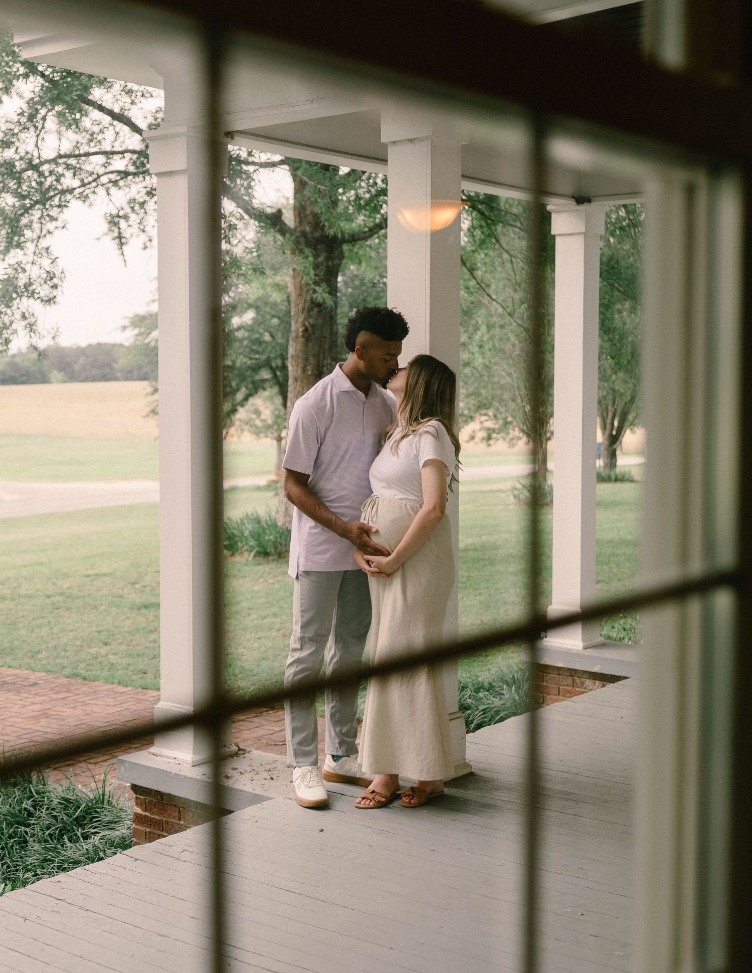 Couple shares a tender kiss on a charming farmhouse porch, framed through a window with garden views.