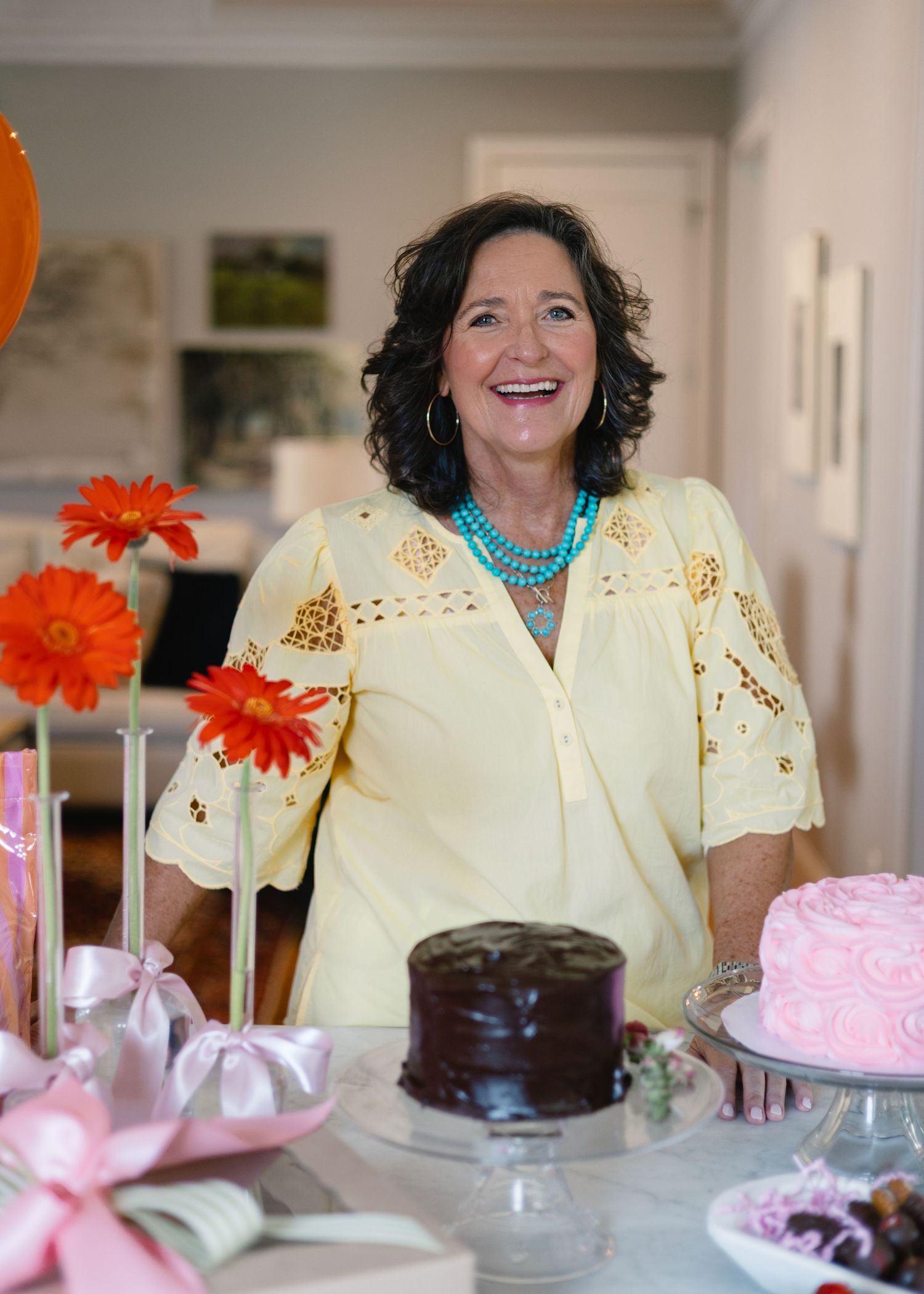 Smiling woman in yellow blouse standing behind a table with cakes and orange flowers.