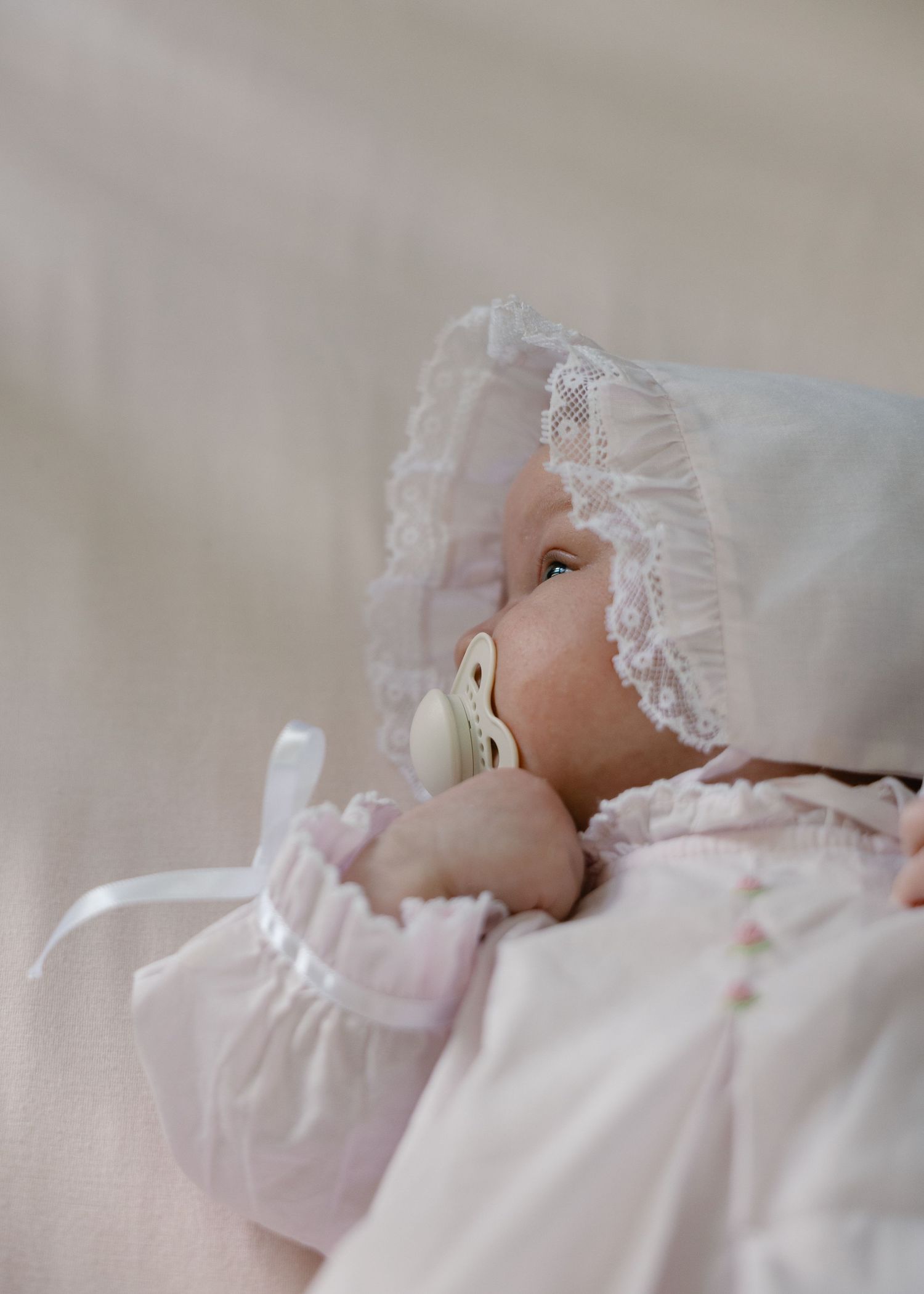 Newborn baby girl in white lace bonnet and pink dress with pacifier.