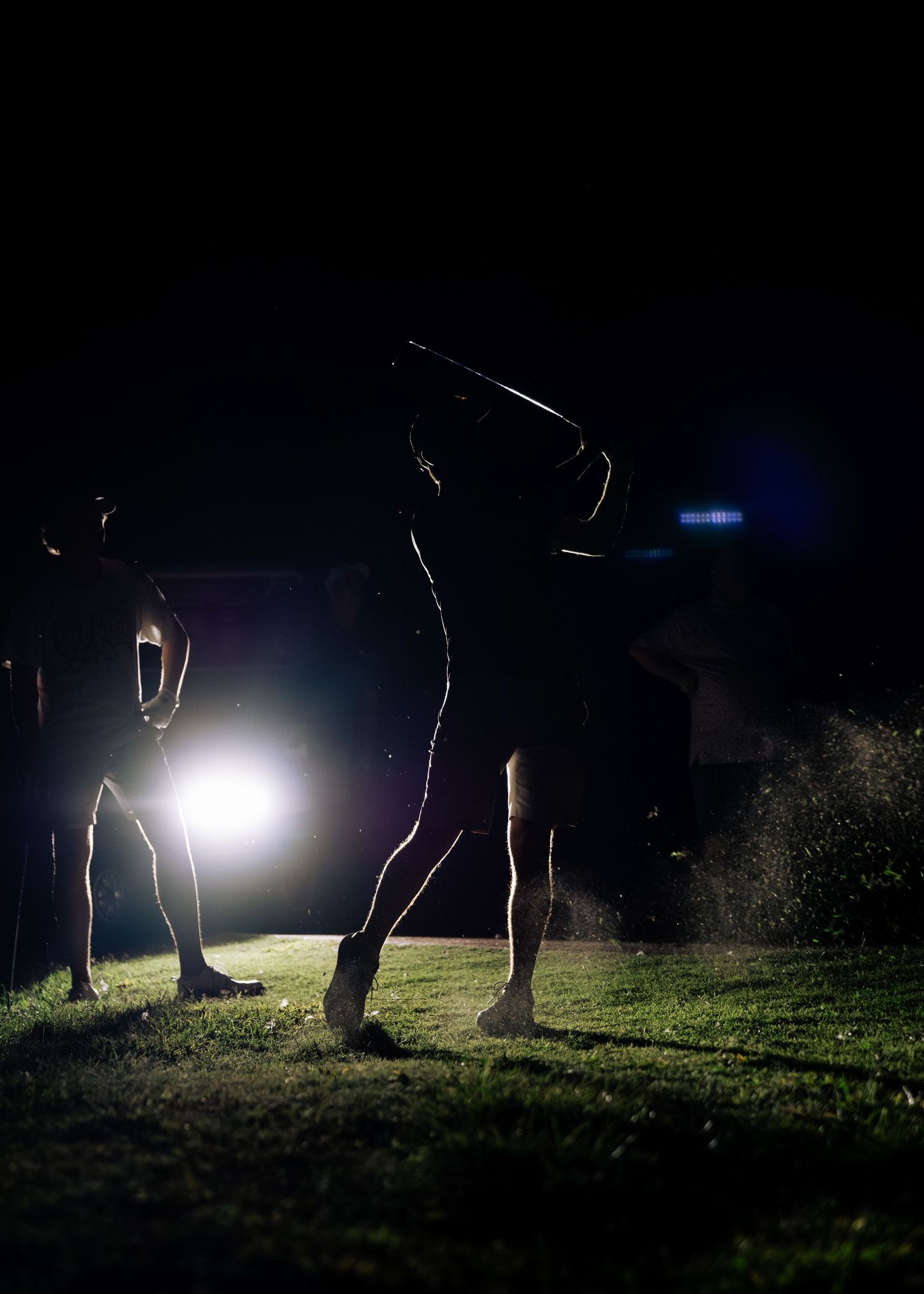 Silhouetted golfers swing clubs under bright spotlight on a dark nighttime golf course.