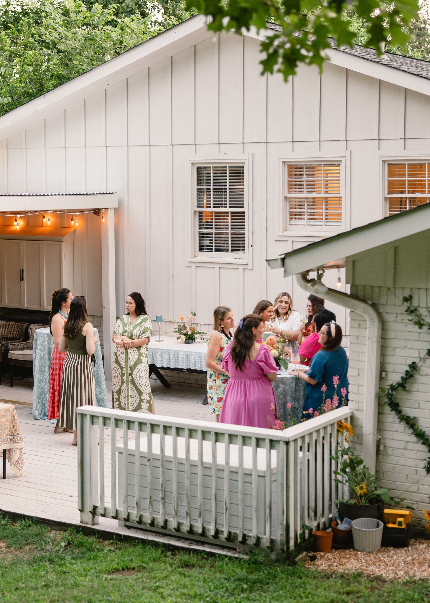 Group of women socializing on a white farmhouse deck during a summer outdoor garden party at dusk.