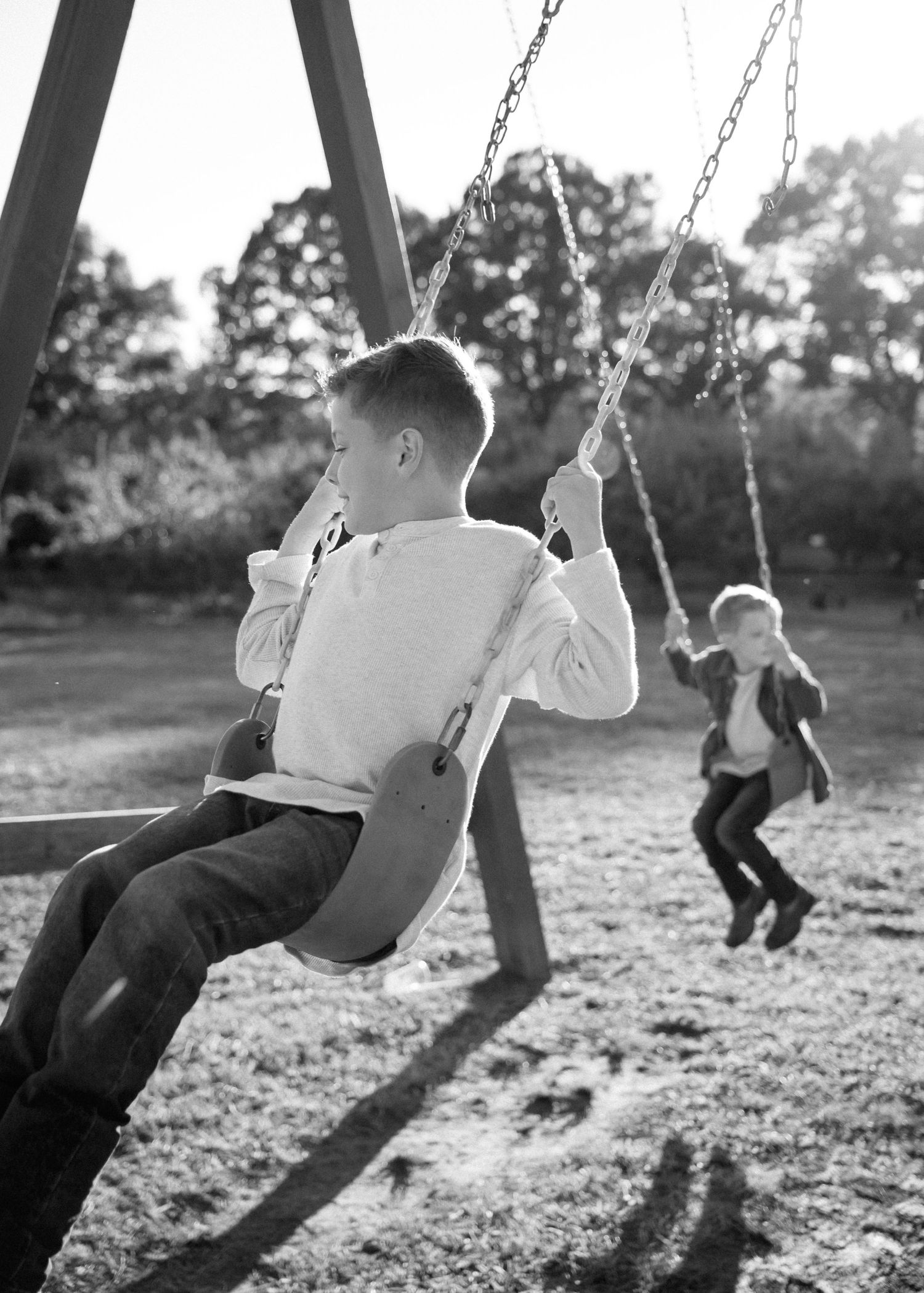 Two boys swing on a playground swing set in a sunlit park with trees in the background, shown in black and white.