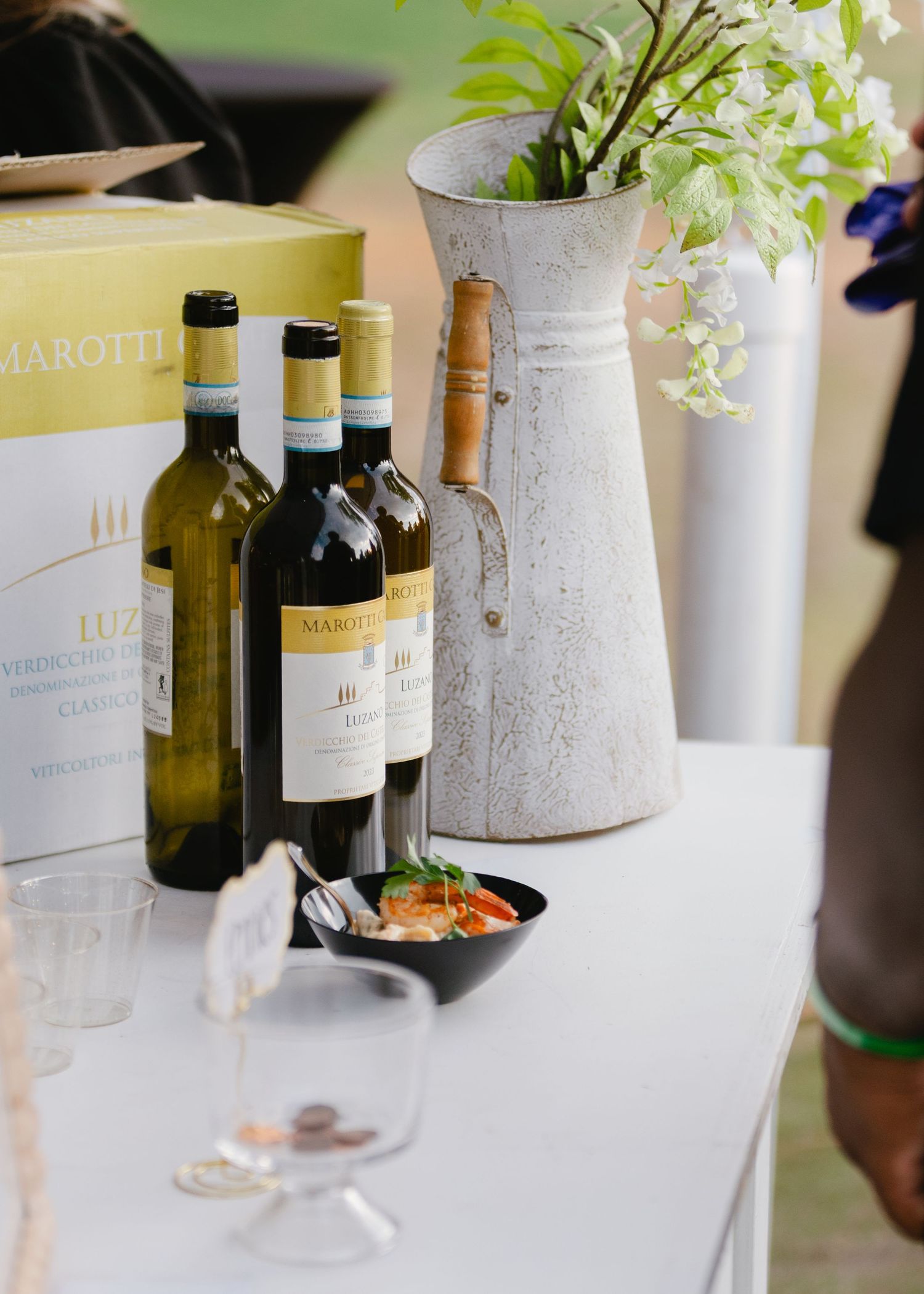 Three Marotti Campi wine bottles on a white table beside a boxed case, a rustic white pitcher with flowers, and a small appetizer bowl.