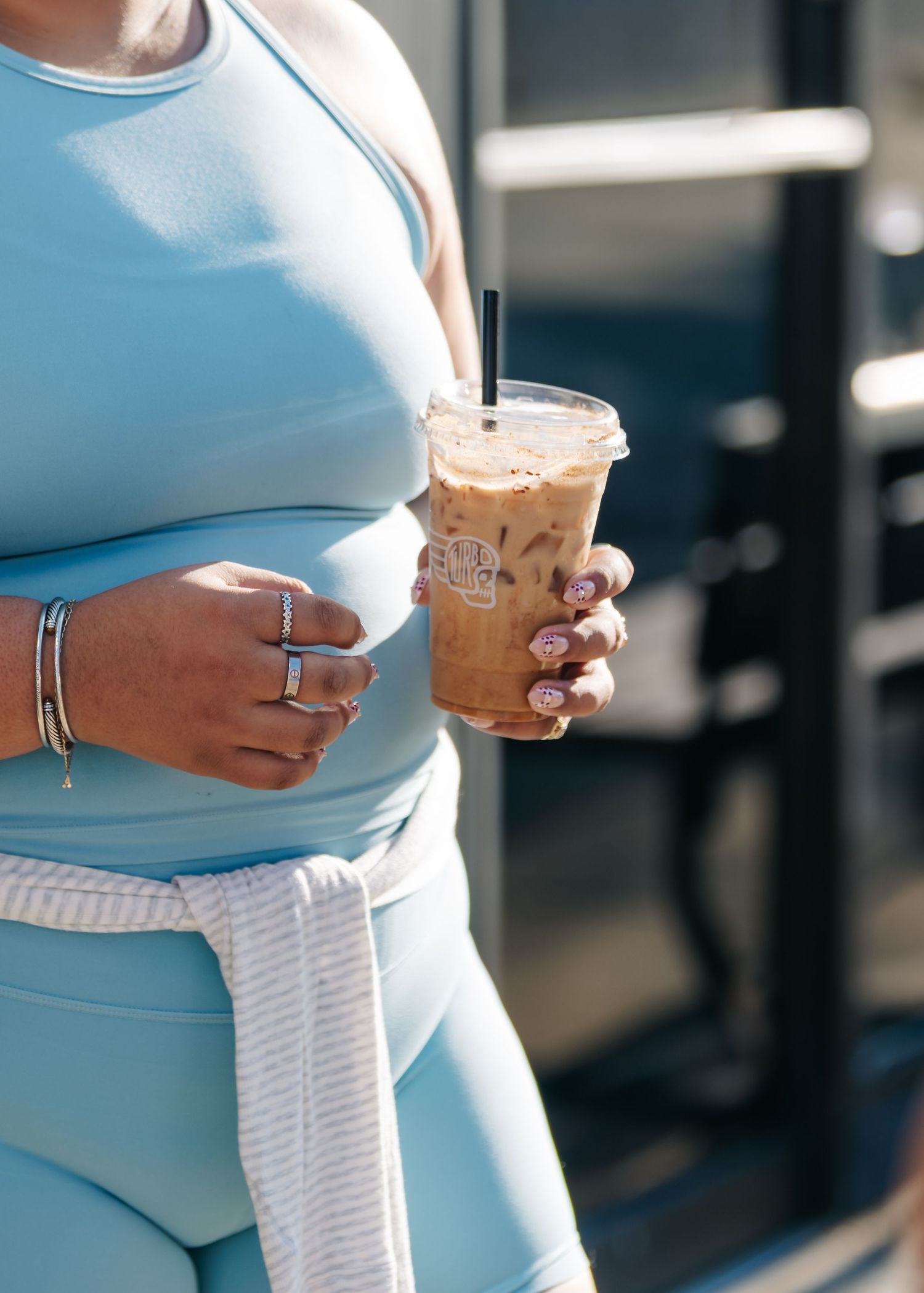 Woman in light blue athletic set holding an iced coffee drink with bracelets and rings on her hand.