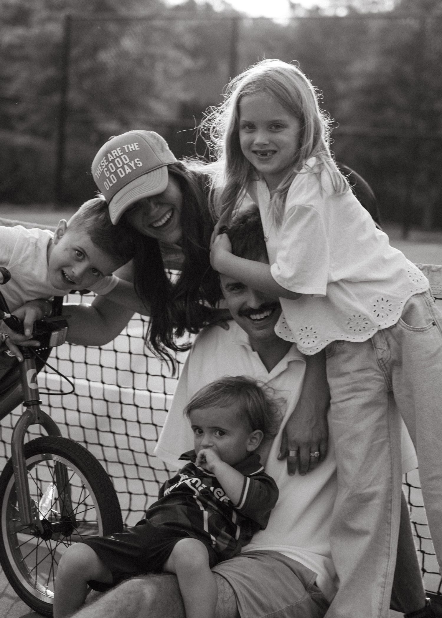 A black and white photo of a joyful family of five laughing together near a tennis net outdoors.