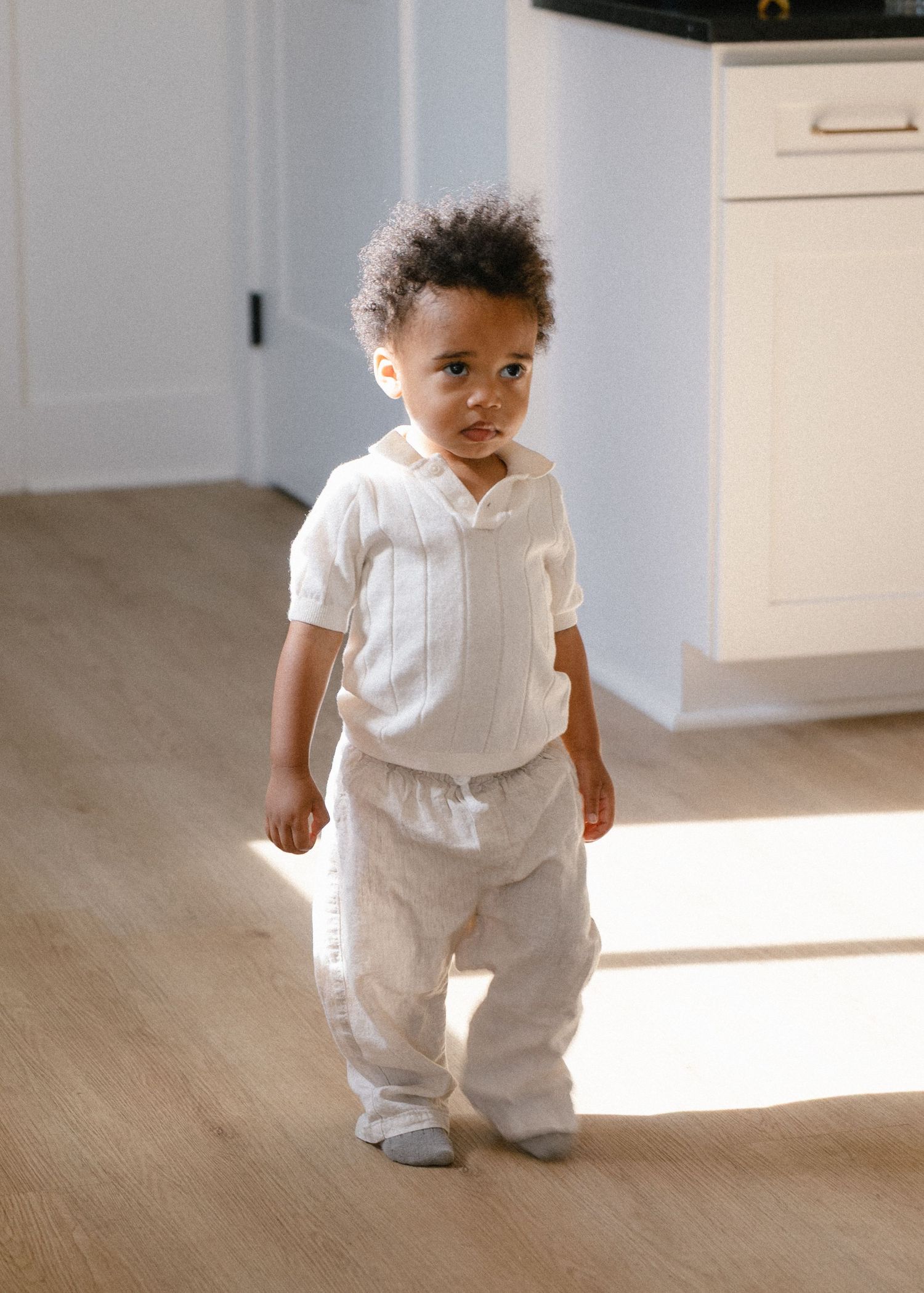 An adorable toddler in a white outfit stands in sunlit kitchen interior.