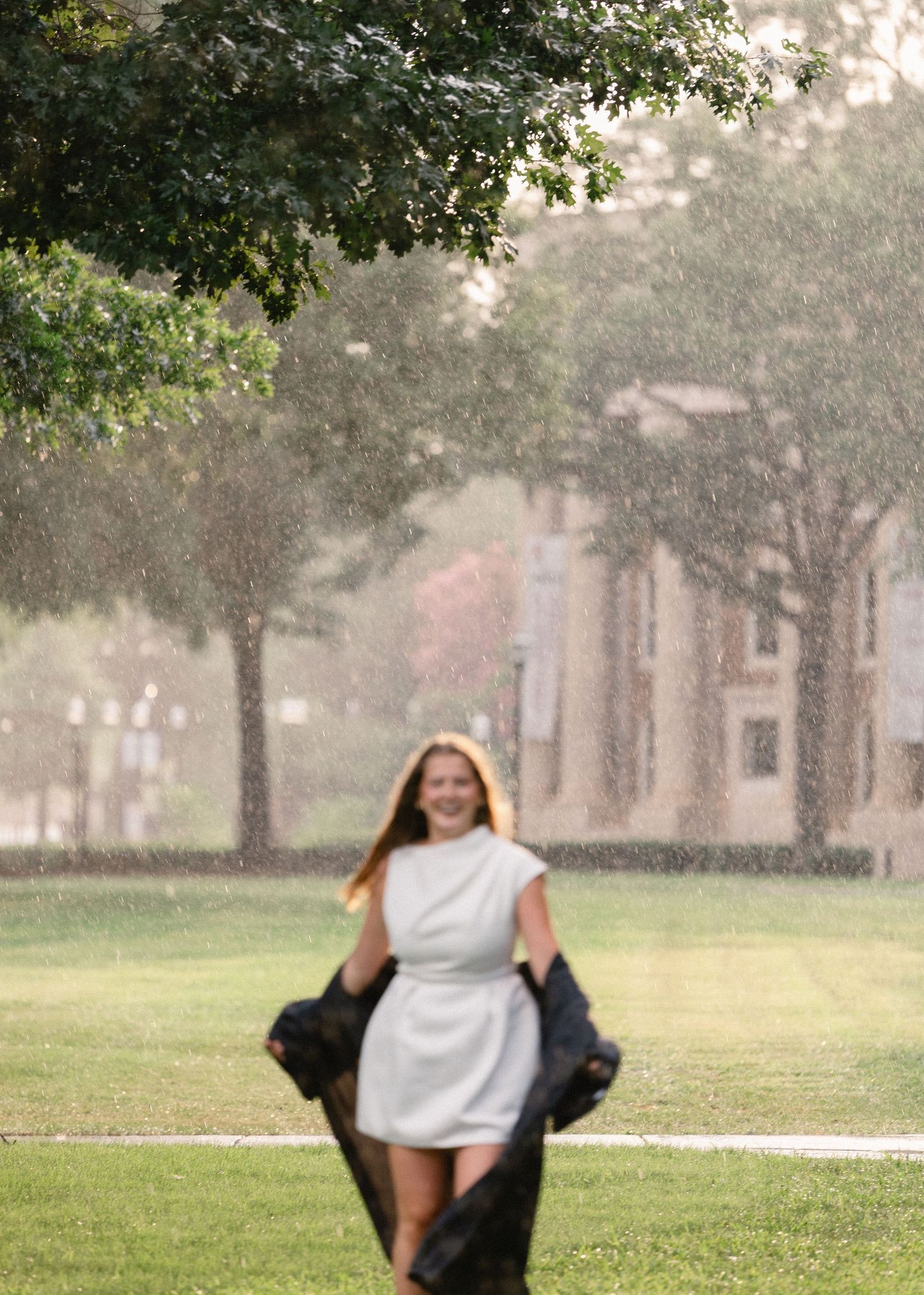 Happy graduate walks through rain in white dress holding black gown on tree-lined campus.