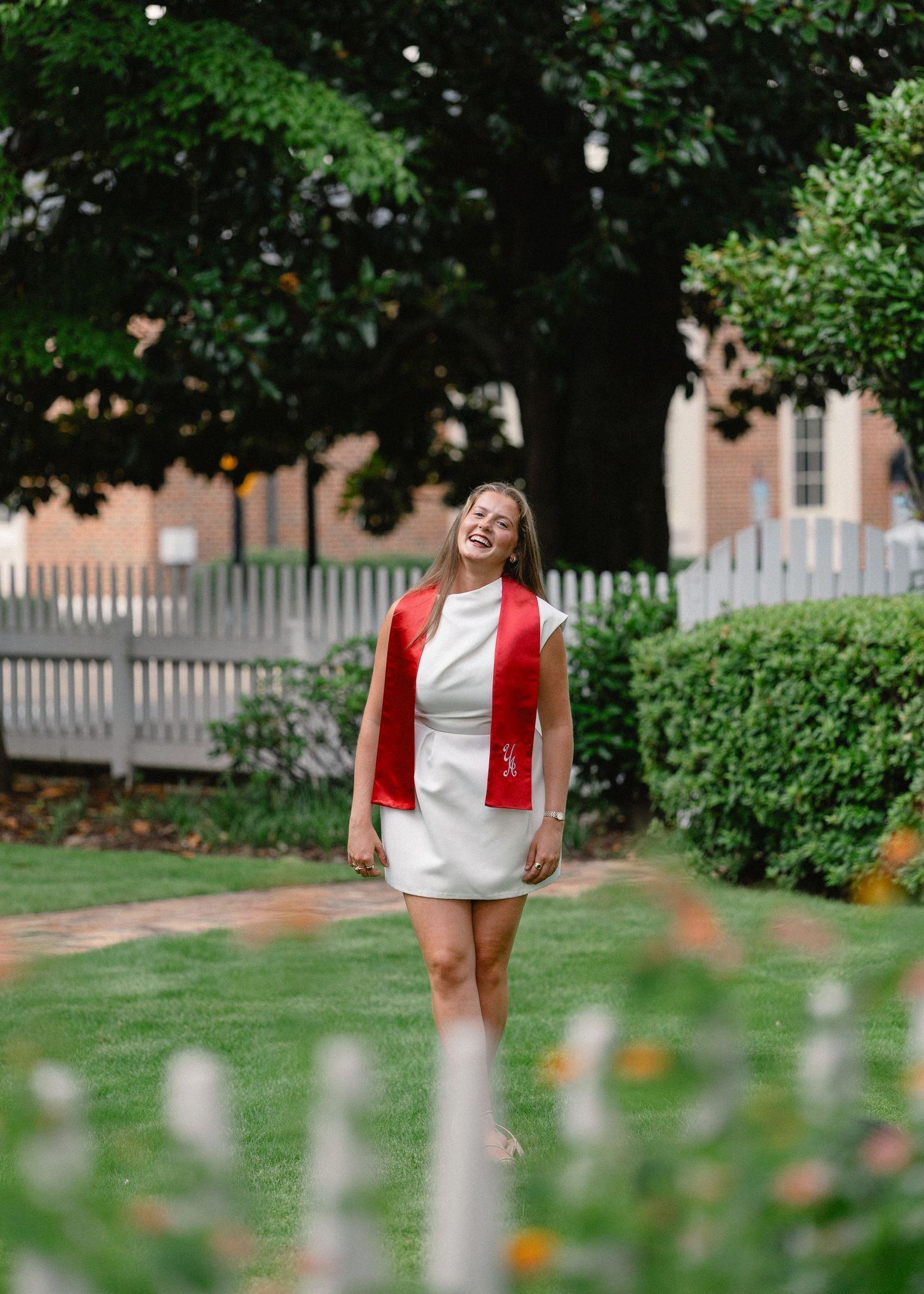 A smiling young woman in a white dress and red graduation sash celebrates outdoors on a sunny day.