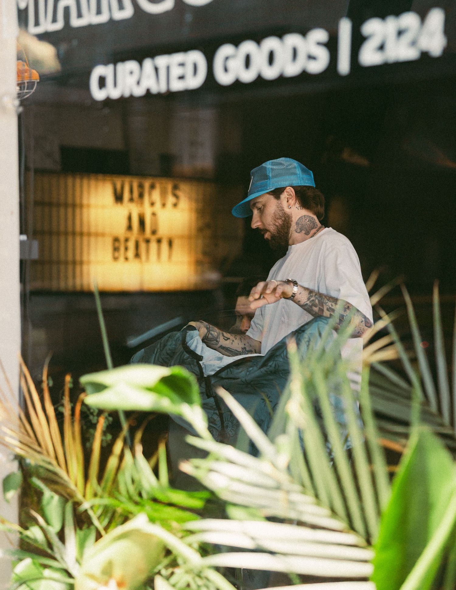 Tattooed man sitting inside Marcus and Beatty curated goods shop at 2124 surrounded by plants.