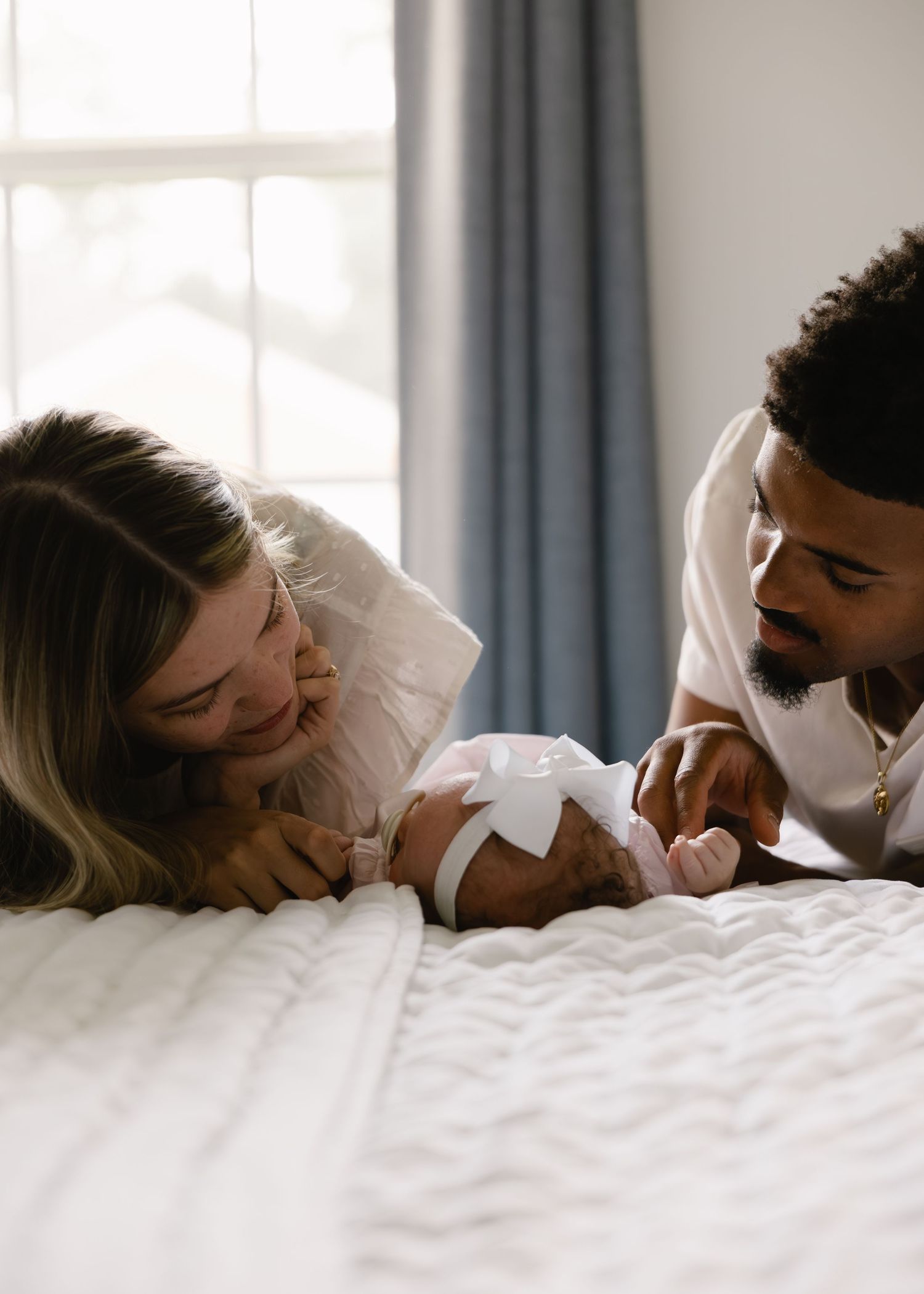 Couple lying on bed smiling at their newborn baby girl with white bow.
