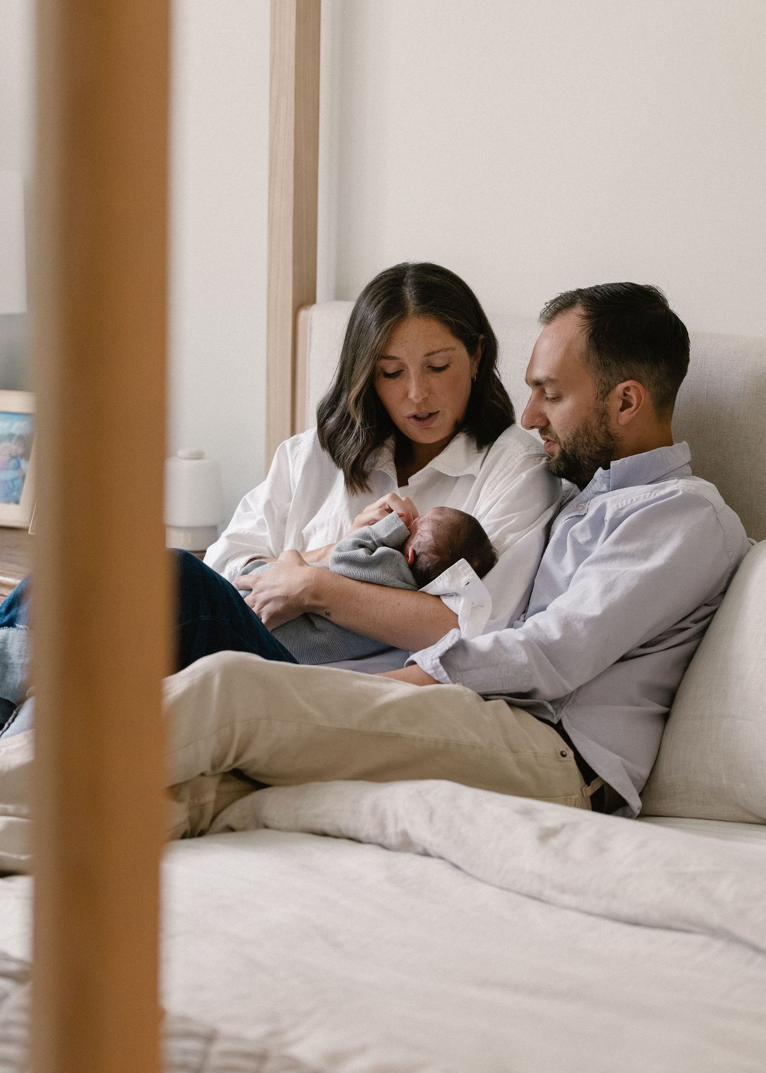 New parents sitting on bed together admiring their newborn baby in a bright cozy bedroom.