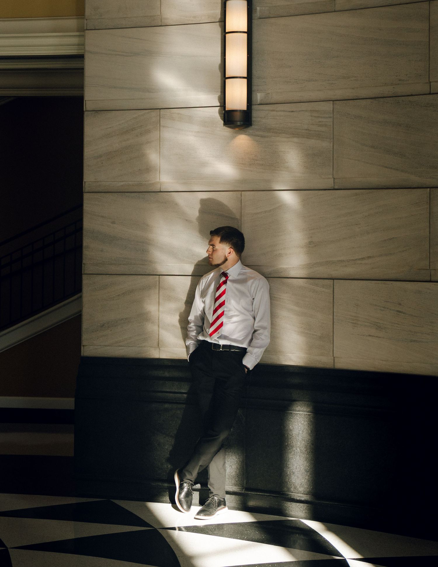 Young man in dress shirt and red tie leaning against marble wall in dramatic light and shadow.