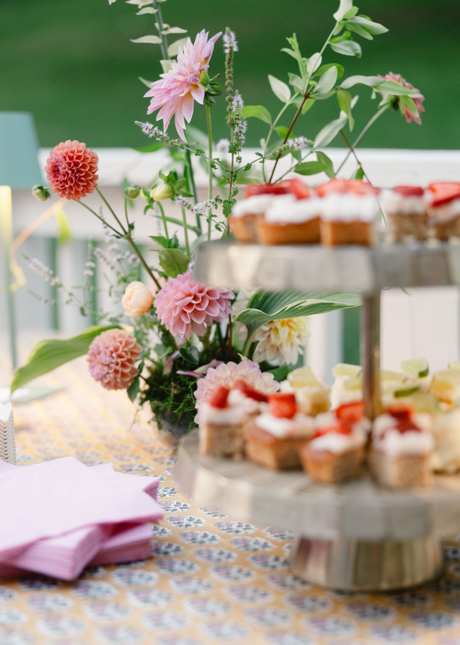 Elegant tiered dessert stand with strawberry topped cakes beside pink dahlia floral arrangement outdoors.