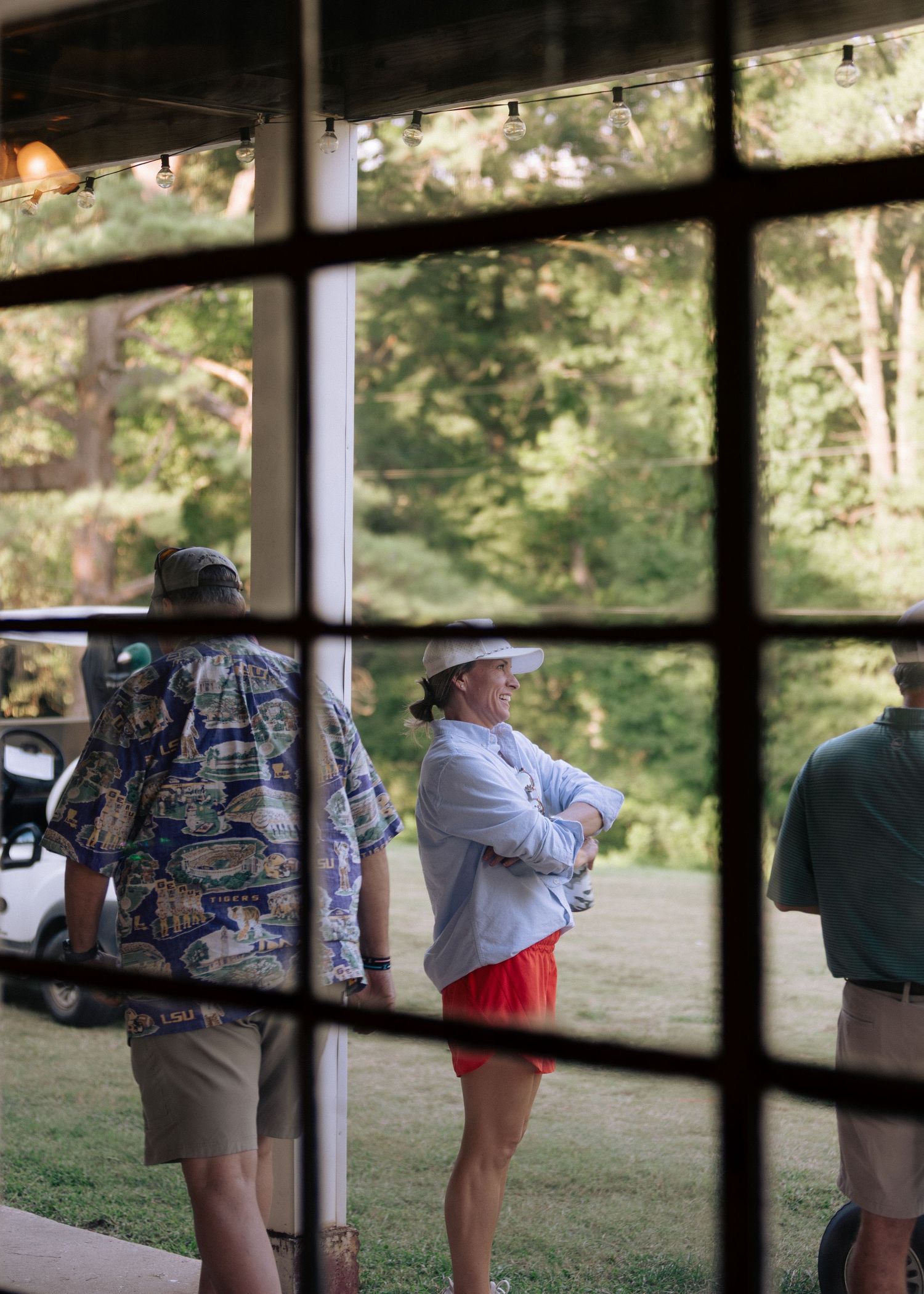 Golfers socializing outside clubhouse viewed through window panes on sunny day.