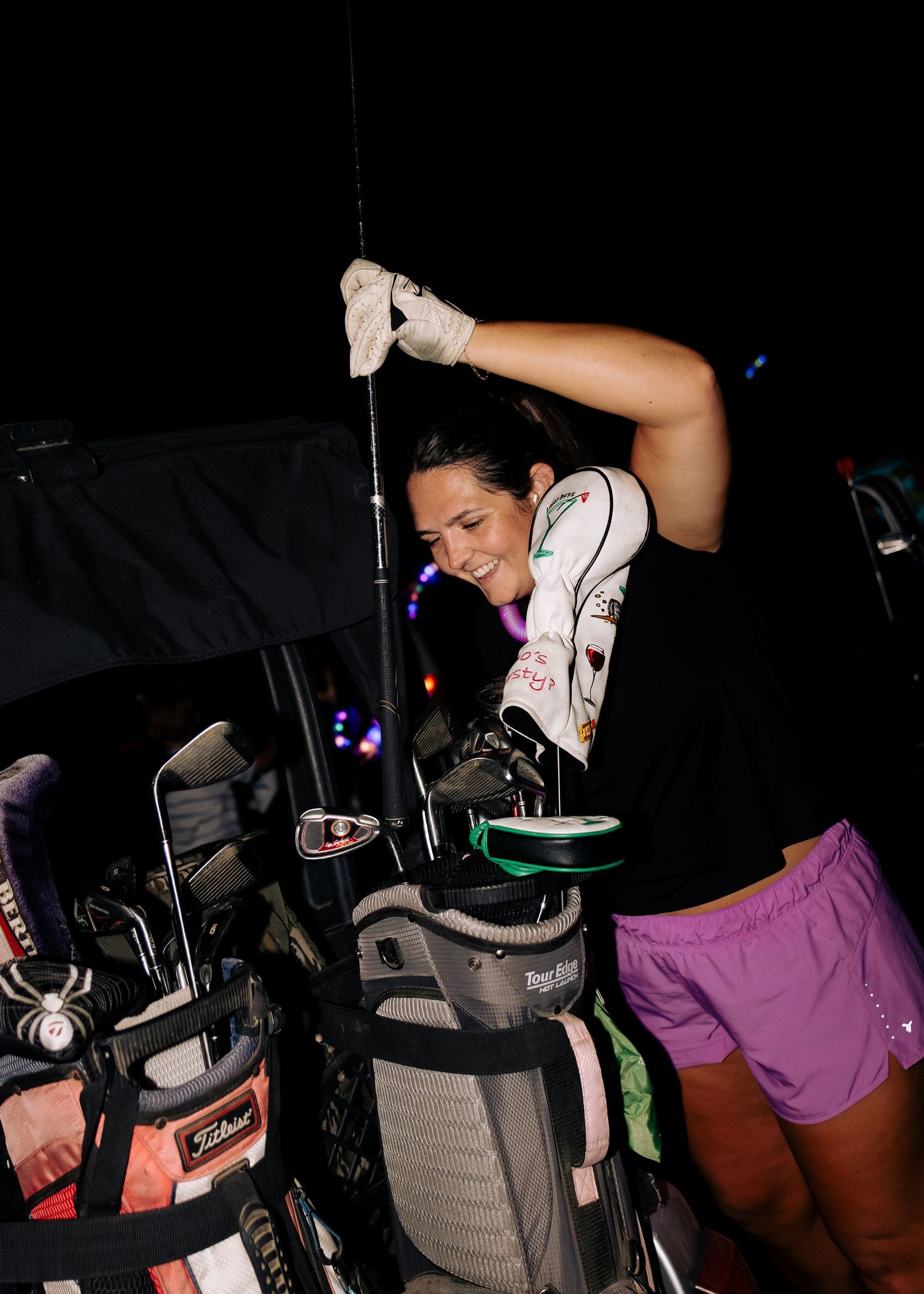 Smiling woman in pink shorts selects a golf club from a bag during a night golf event.